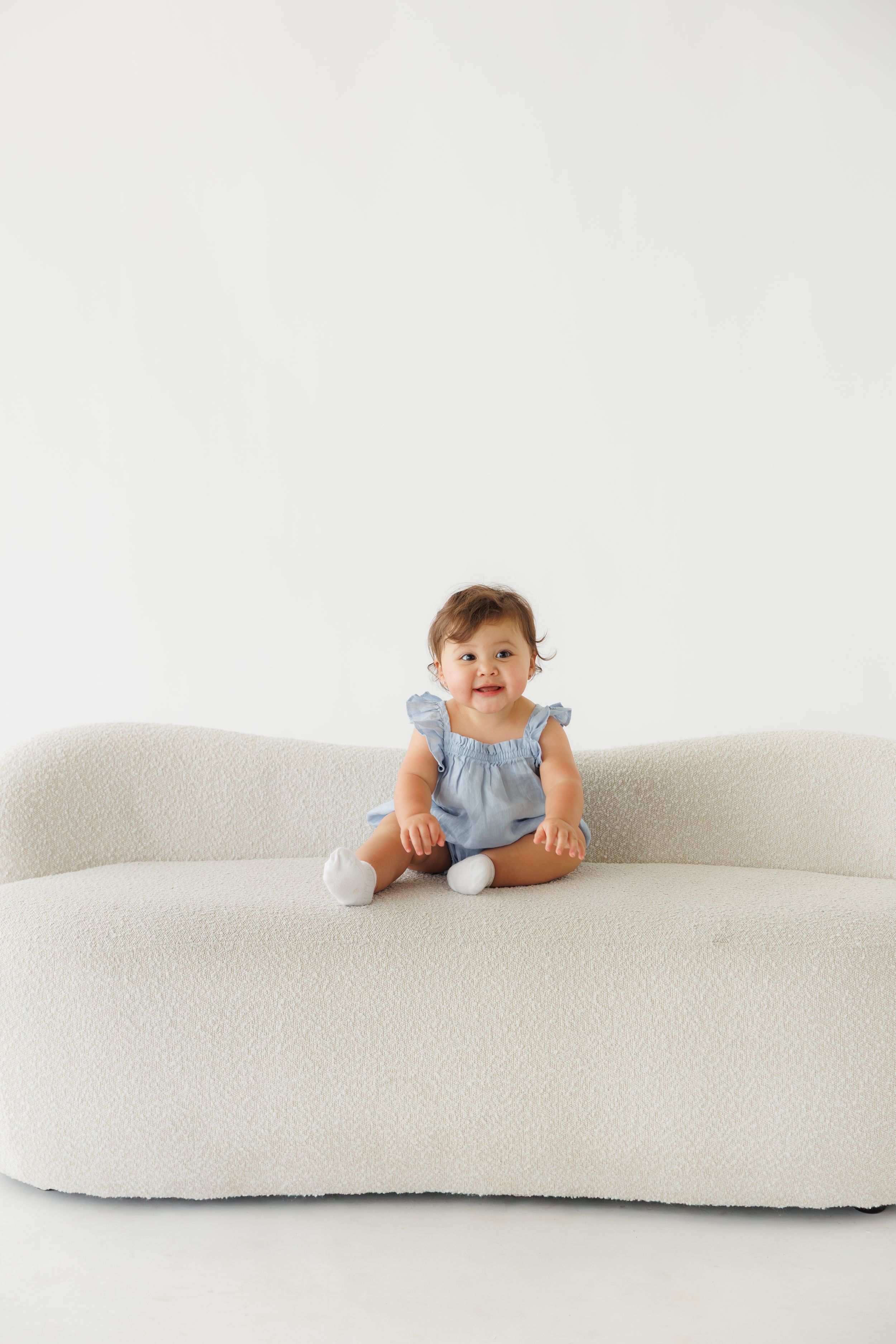 Smiling baby girl in blue romper sitting on a white boucle sofa against a white background.