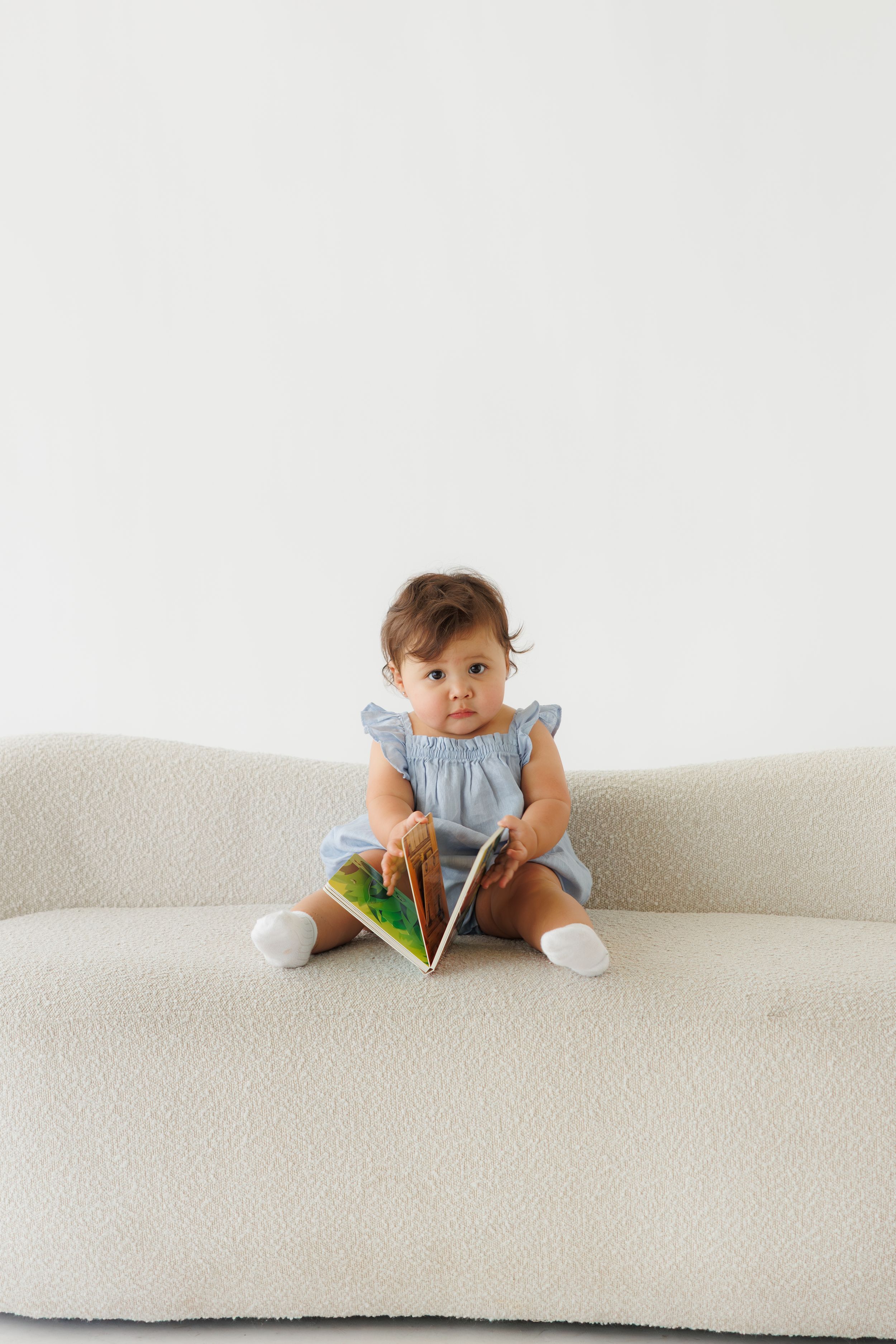 A baby girl in a blue dress sits on a cream sofa, holding a colorful book and looking at the camera.
