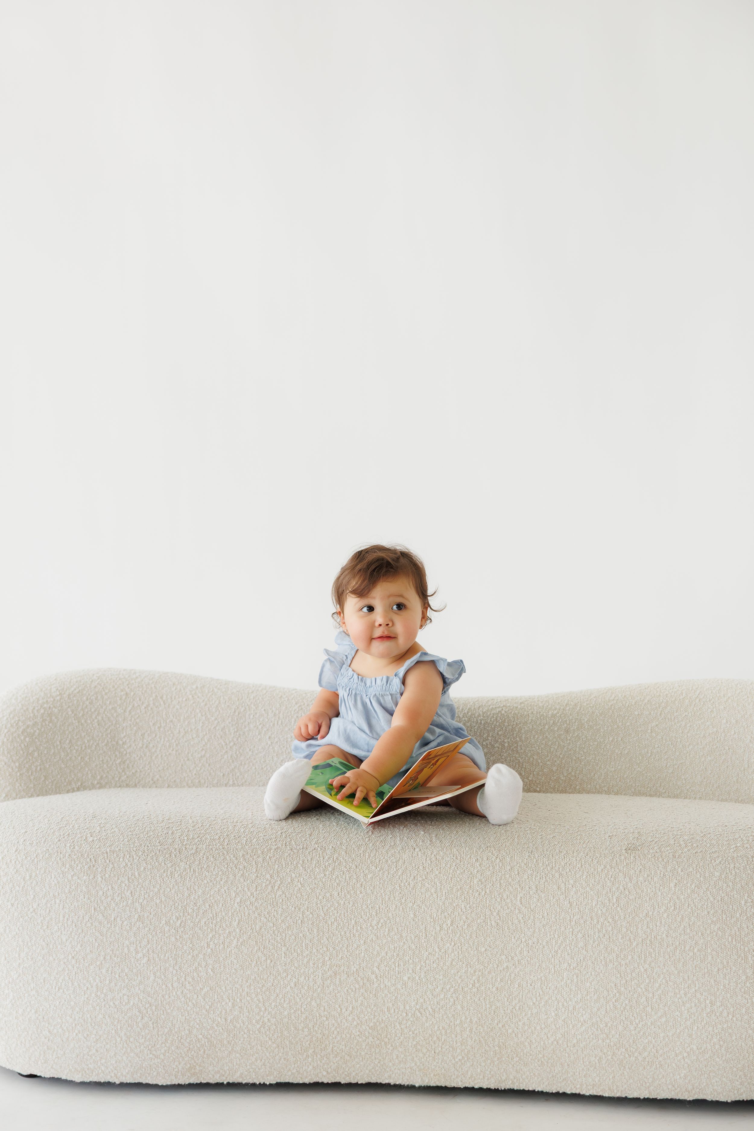 A baby girl in a blue dress sits cross-legged on a cream boucle sofa, holding a colorful book.