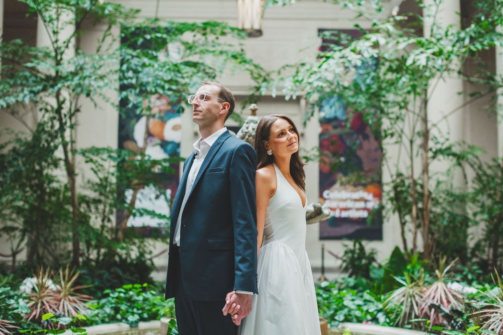 Bride and groom holding hands back-to-back in a lush indoor garden atrium on their wedding day.