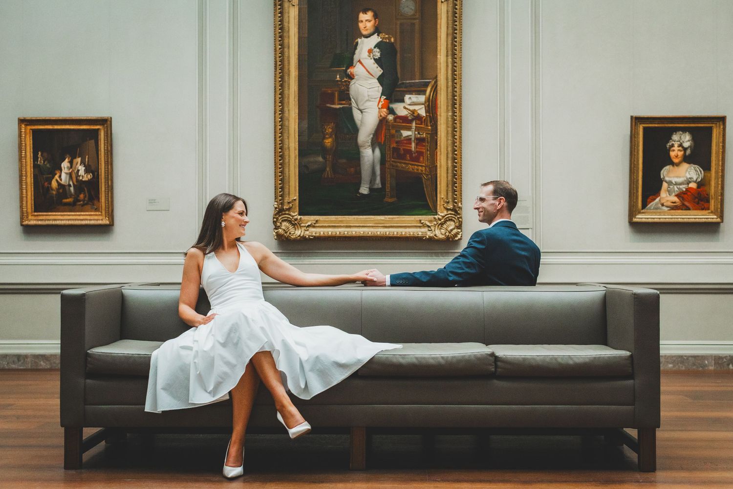 Couple in wedding attire sitting on a modern sofa in an art museum with classic paintings on the wall.