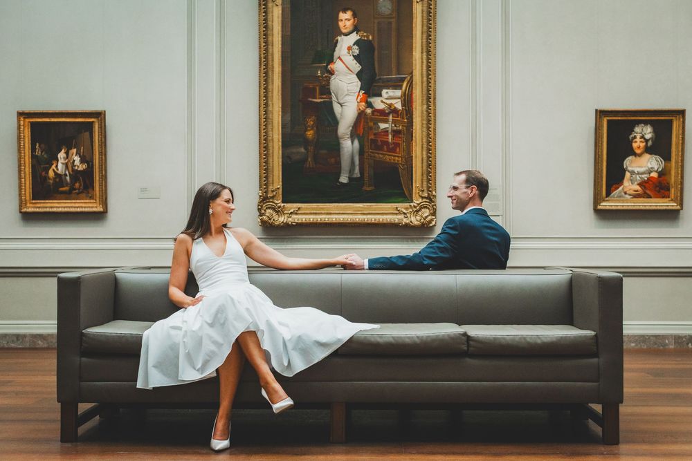 Couple in wedding attire sitting on a modern sofa in an art museum with classic paintings on the wall.
