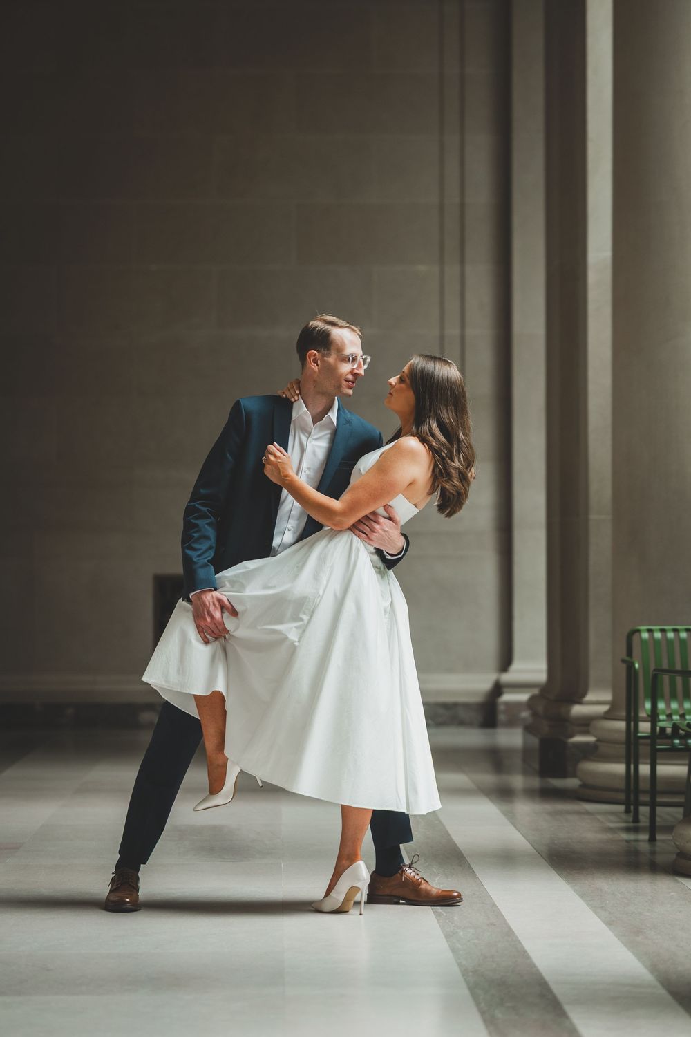 Couple dancing elegantly in a grand hall, groom in navy suit, bride in white midi dress and heels, sharing a joyful moment.