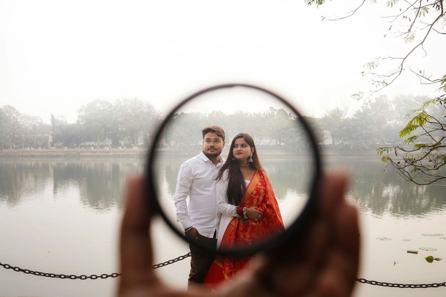 Couple posing romantically by a misty lake, framed through a circular lens, man in white shirt, woman in red saree.