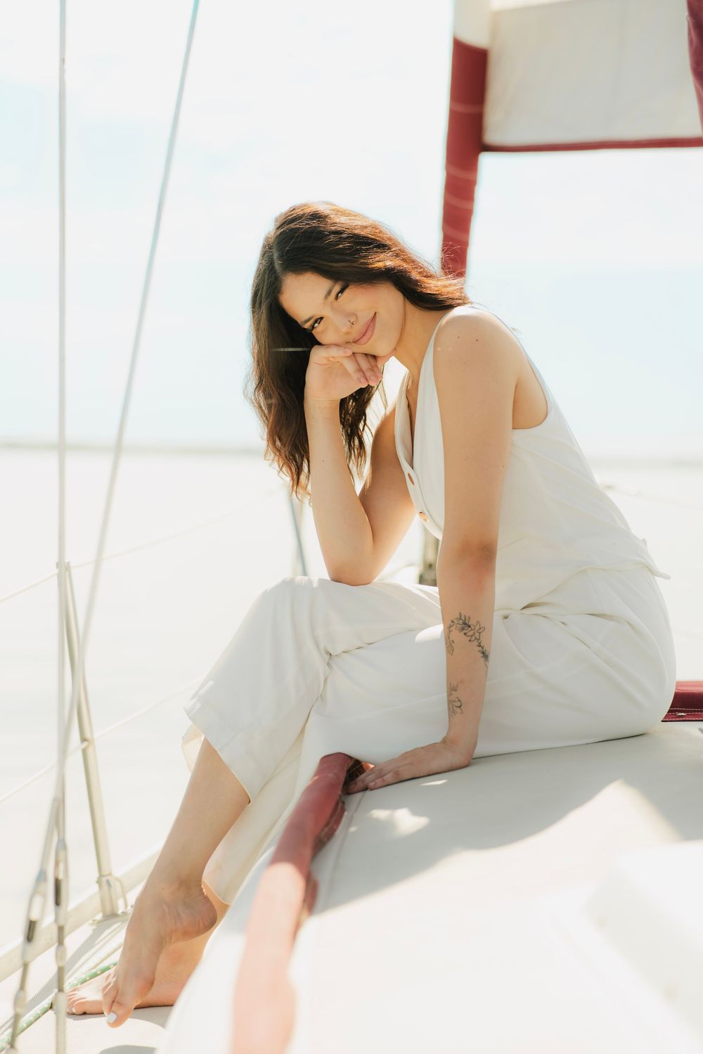 A smiling woman with a tattoo sits relaxed on a sailboat deck, wearing white linen outfit, enjoying a sunny day at sea.