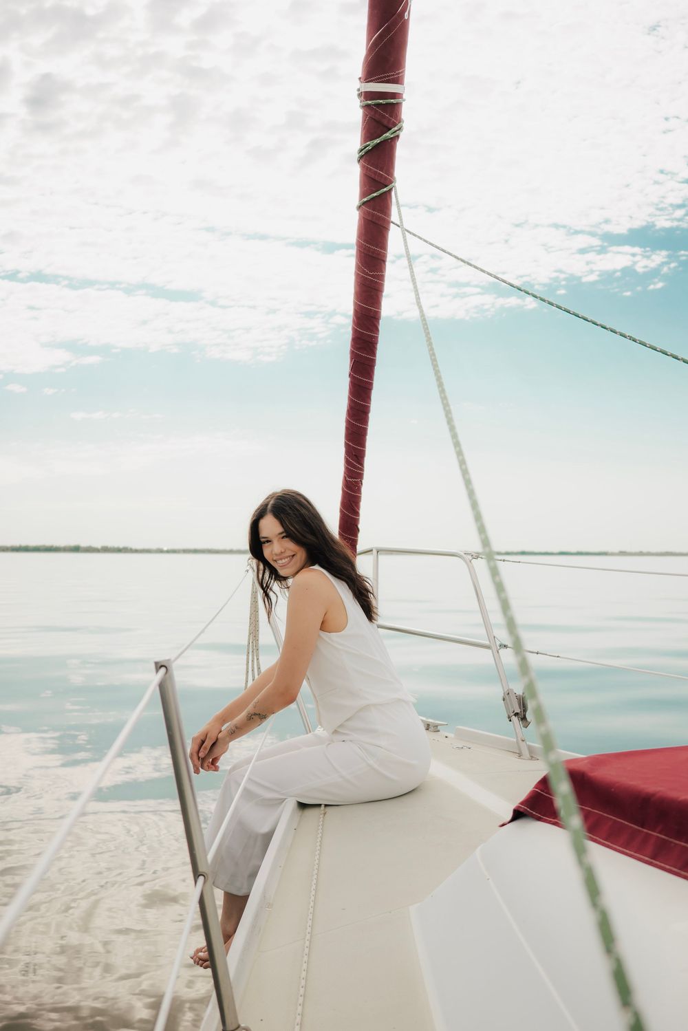 Smiling woman in white outfit sitting on sailboat bow with calm water and cloudy sky backdrop.