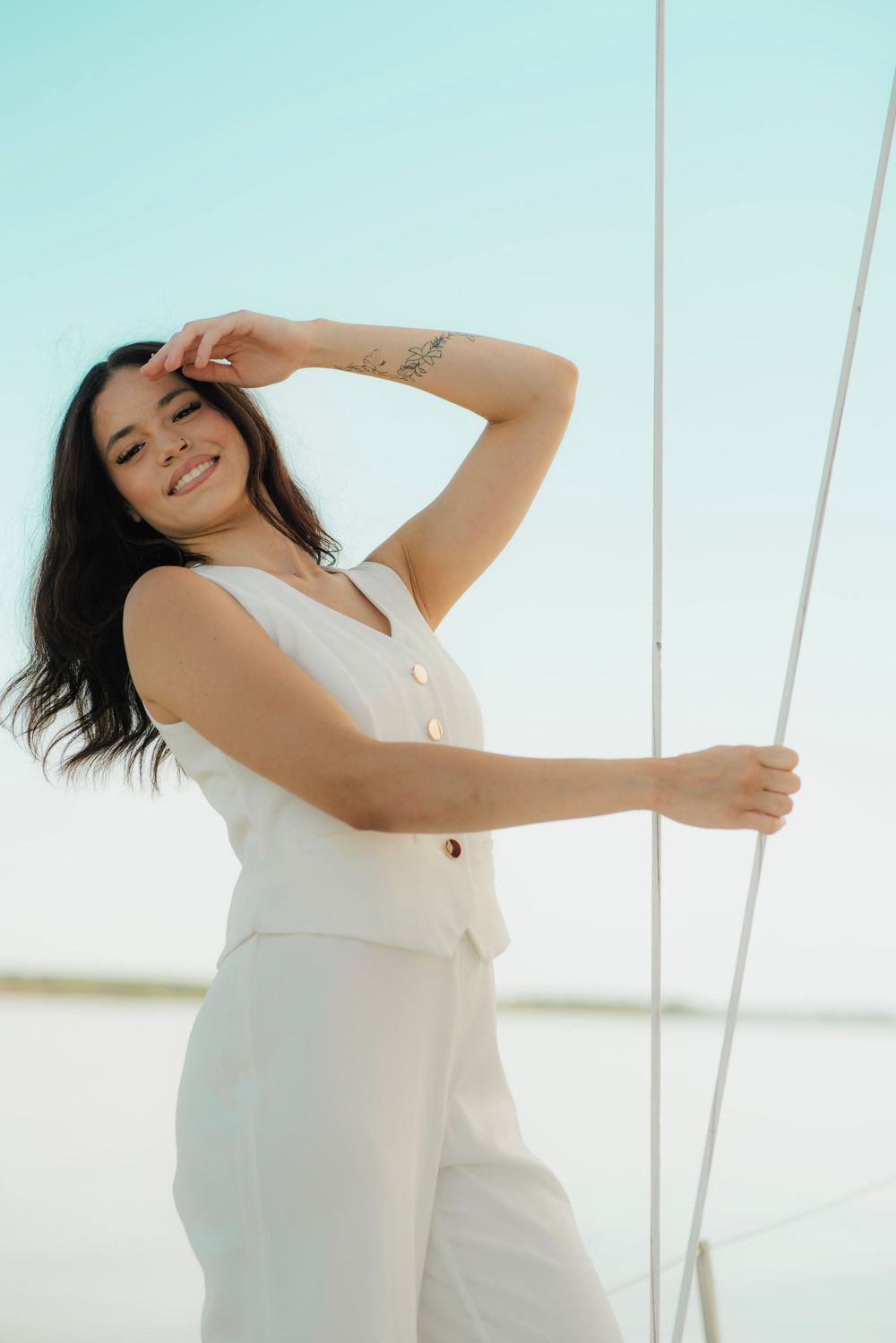 Smiling brunette woman in white pantsuit posing by sailboat mast under blue sky.