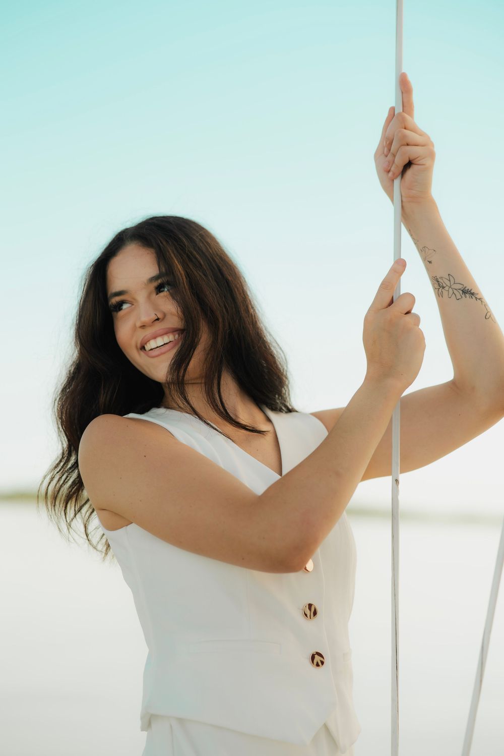 Smiling young woman in white sleeveless outfit holding a rope outdoors with a clear blue sky background.