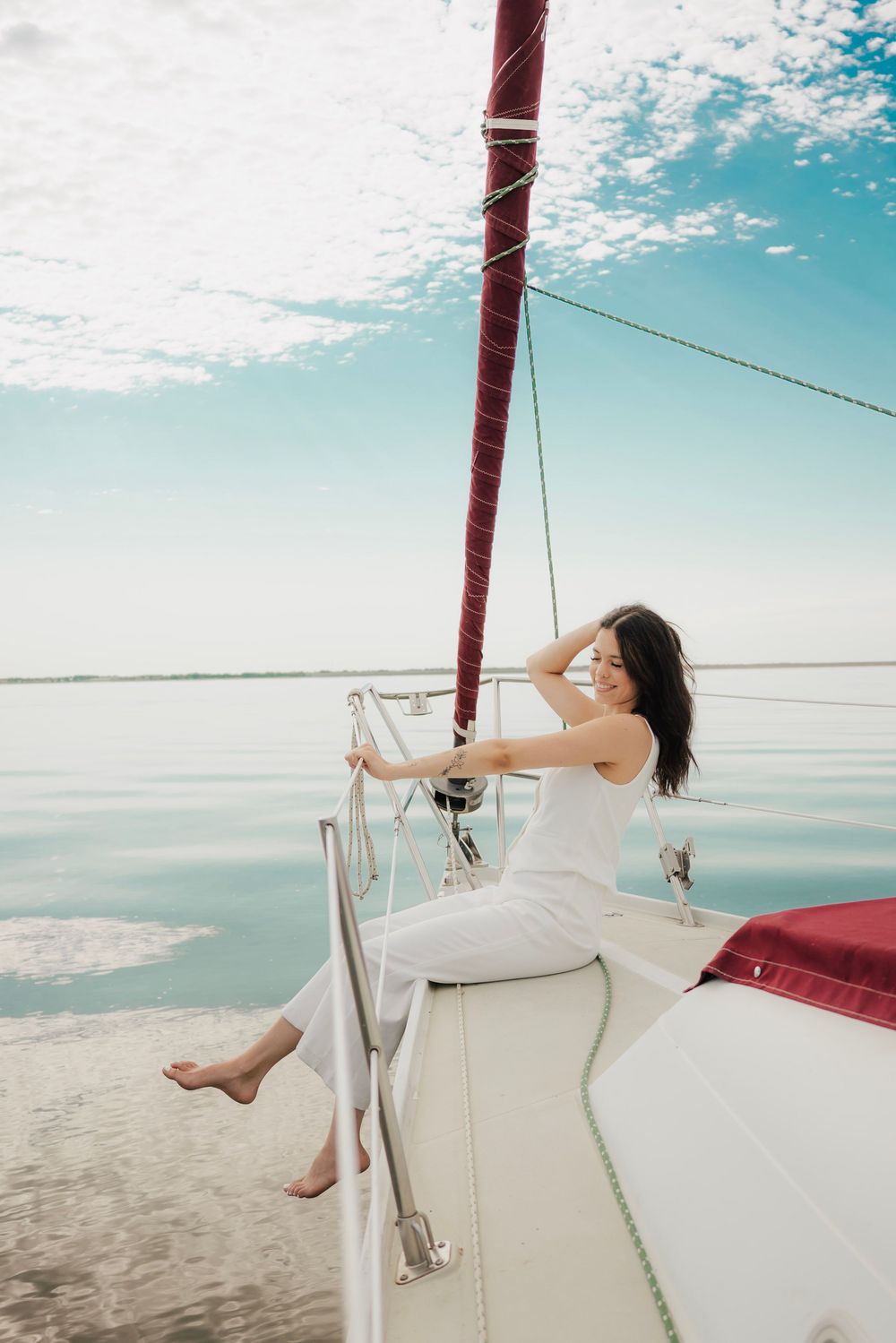 A smiling woman in white outfit relaxes on the deck of a sailboat on calm blue waters under a partly cloudy sky.