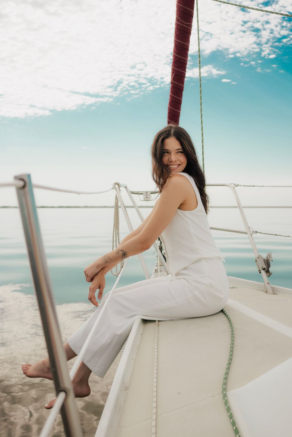 Smiling brunette woman in white outfit sitting on sailboat bow with blue sky behind her.