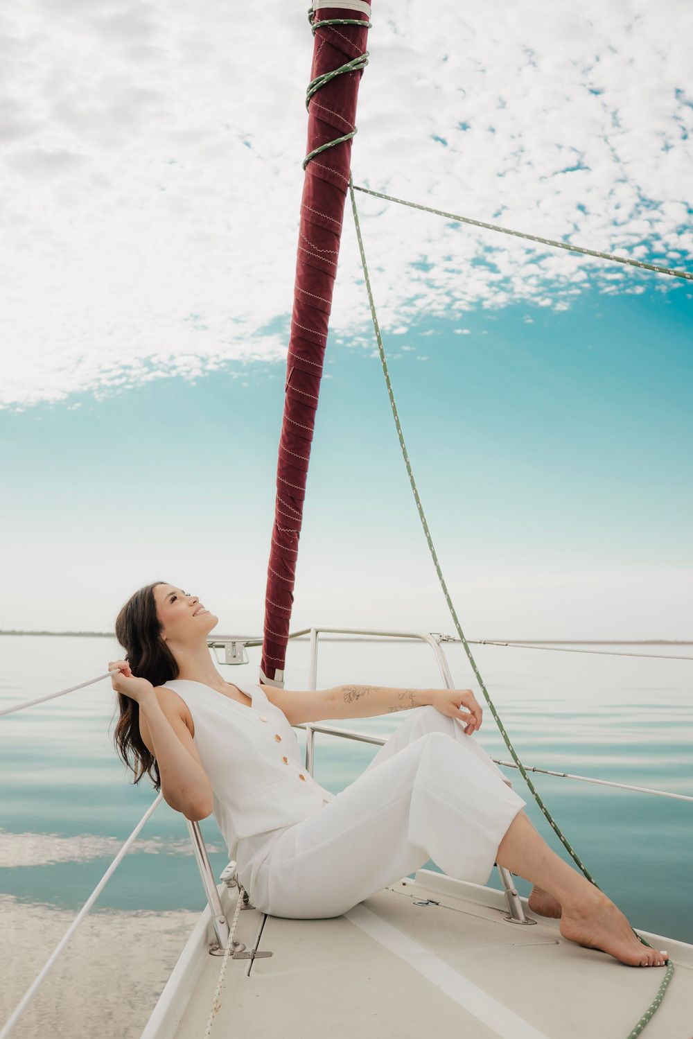 Woman in white outfit relaxing on sailboat bow with calm turquoise sea and cloudy sky backdrop.