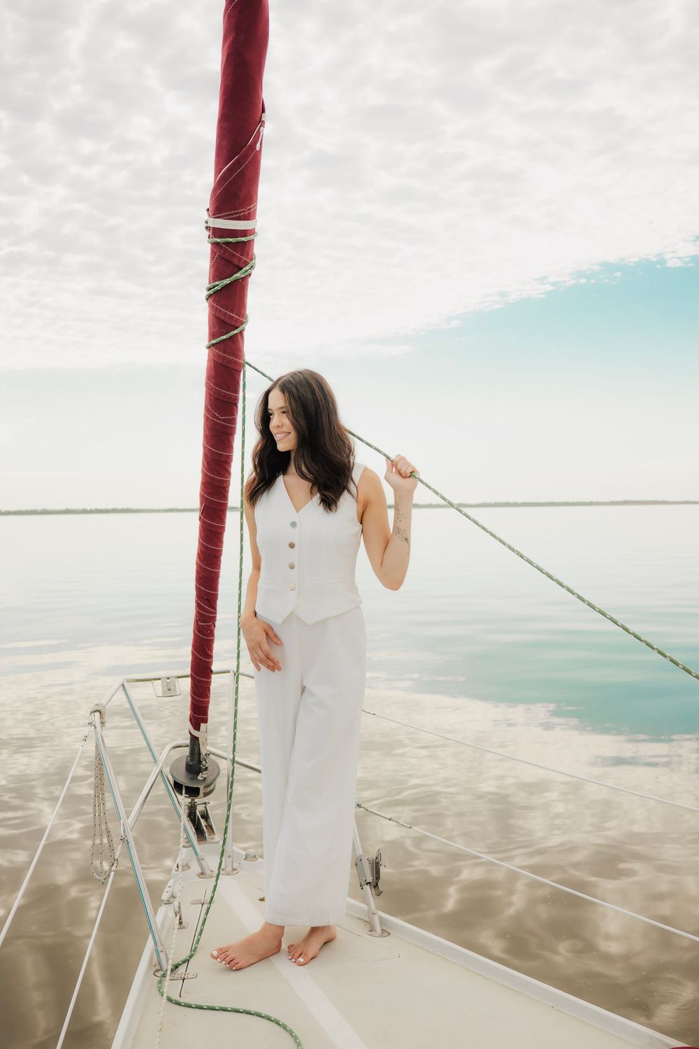 Woman in white outfit standing on sailboat bow holding mast ropes over calm water.