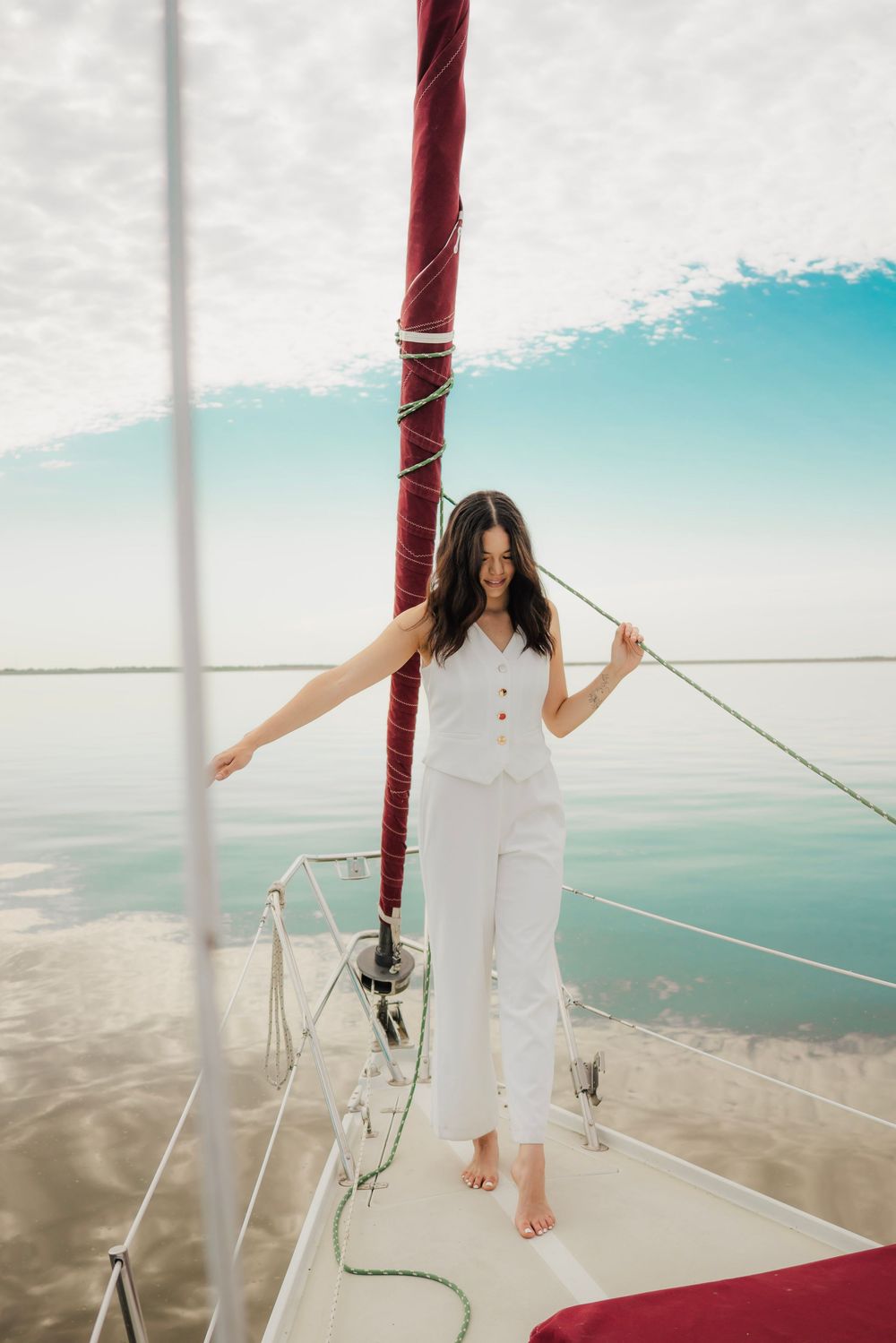 A woman in white outfit stands barefoot on a sailboat deck, holding rigging with calm blue water behind her.