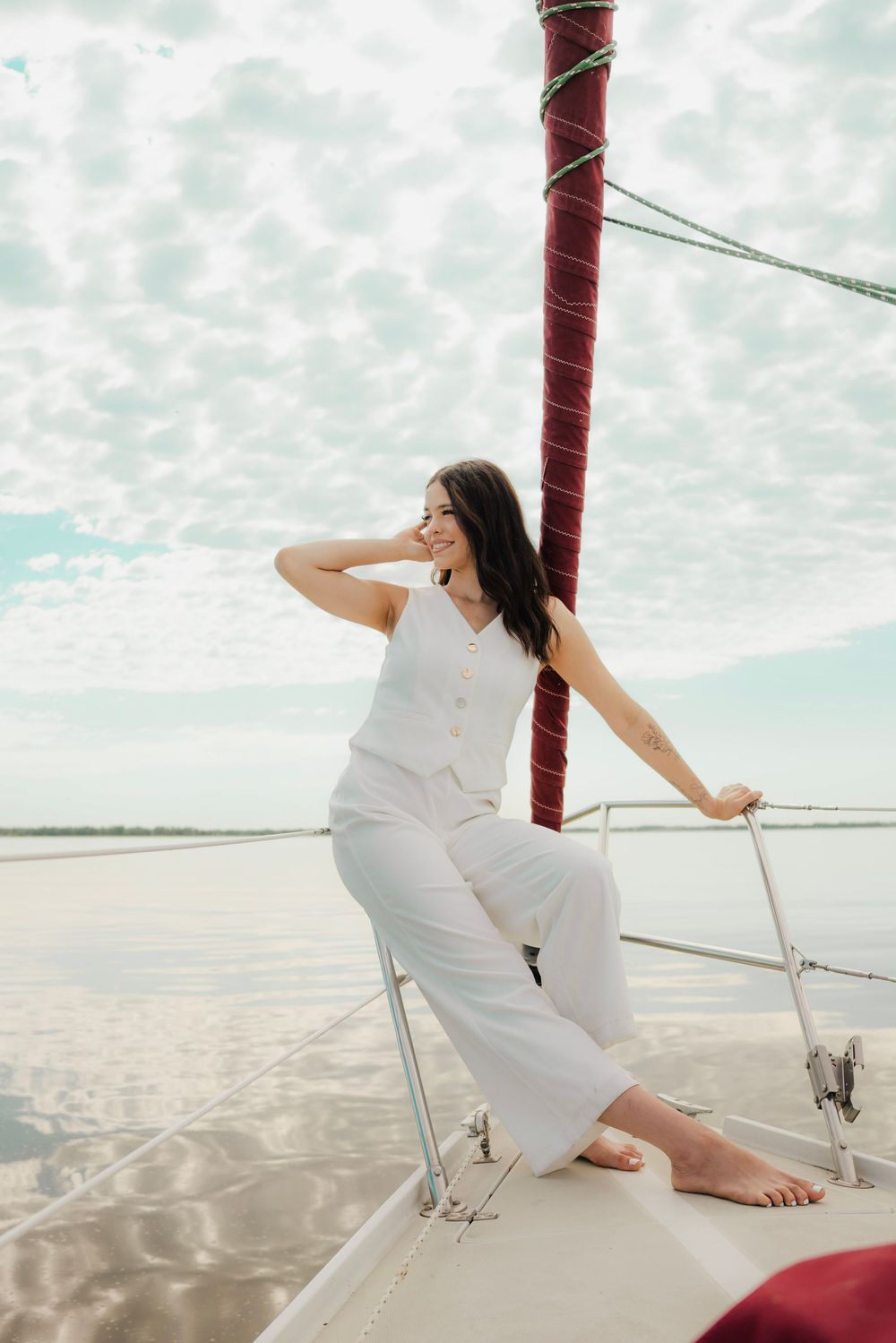 Smiling woman in white ensemble sitting on sailboat bow near mast on calm water.