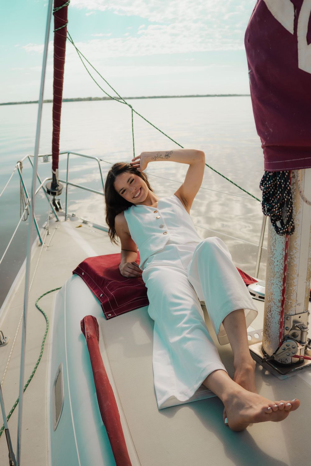 Happy woman in white jumpsuit lounging on sailboat deck laughing with ocean in background.