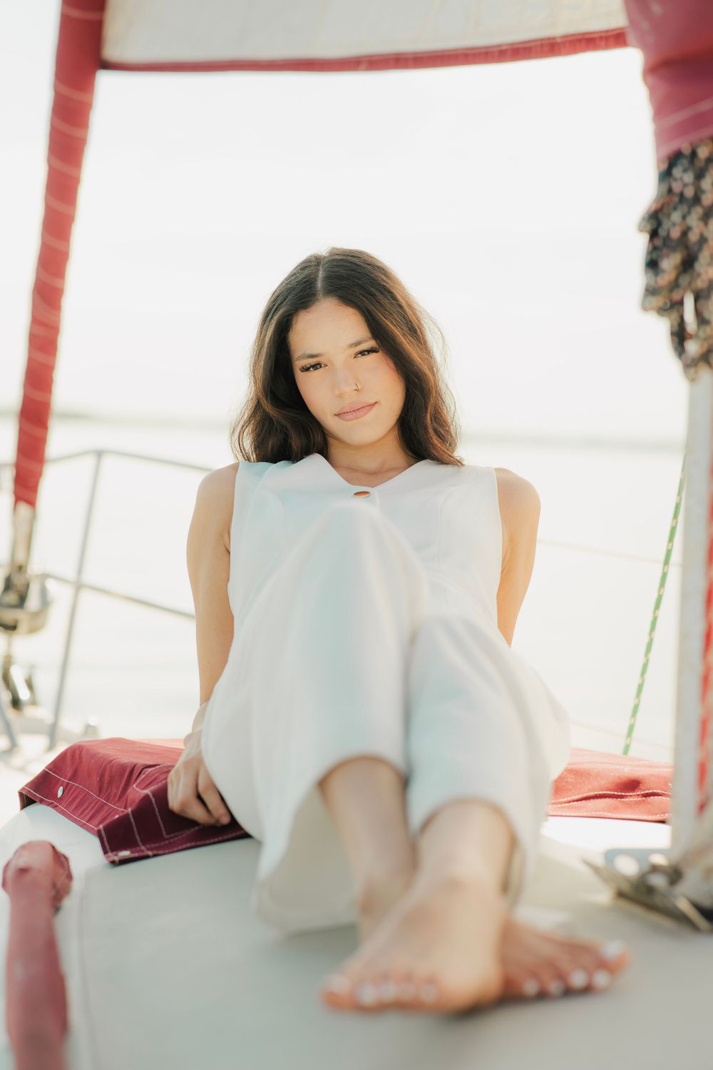 Young woman in white outfit relaxing barefoot on sailboat deck with red accents.