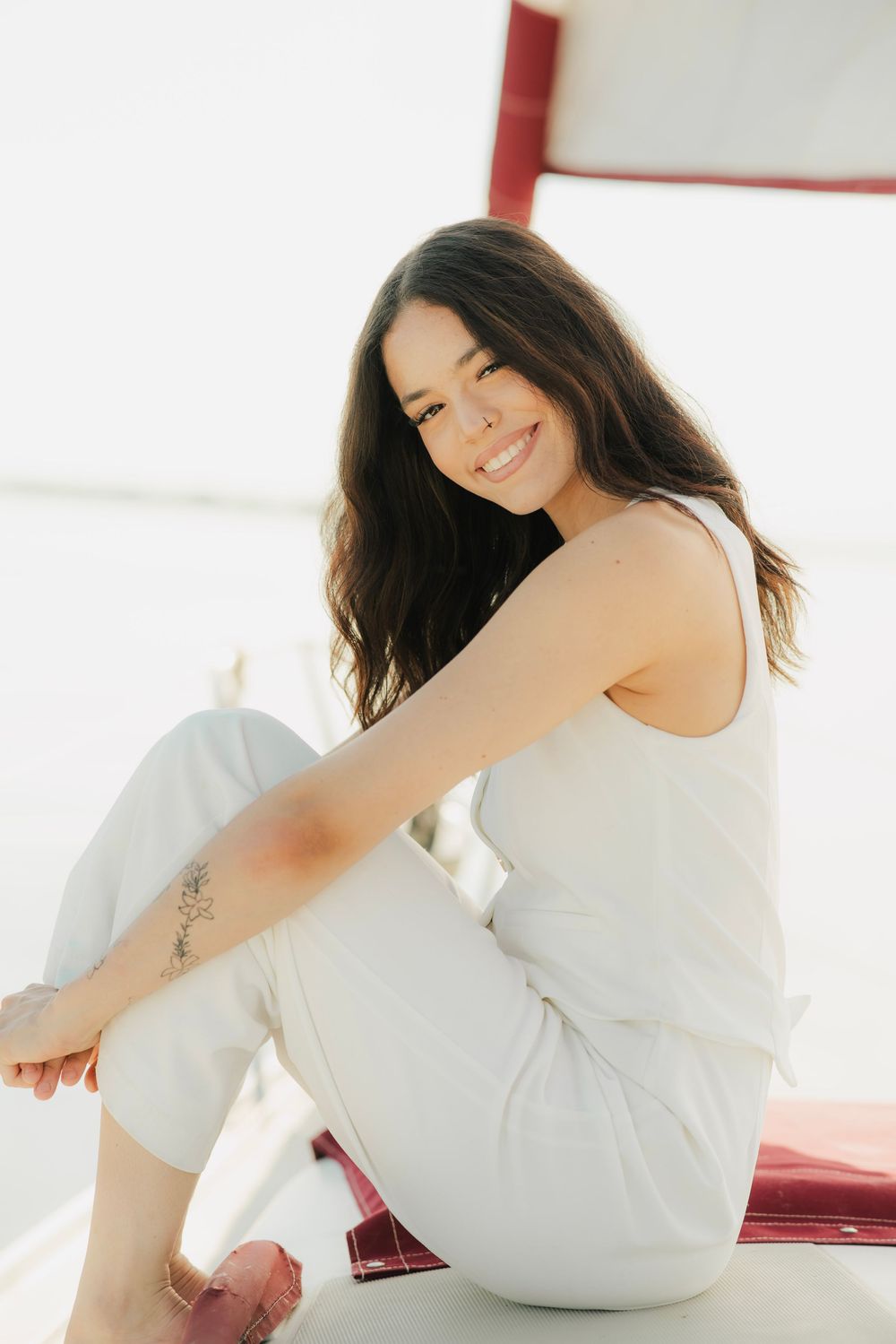 Smiling brunette woman in white outfit with floral tattoo sitting on sailboat in bright light.