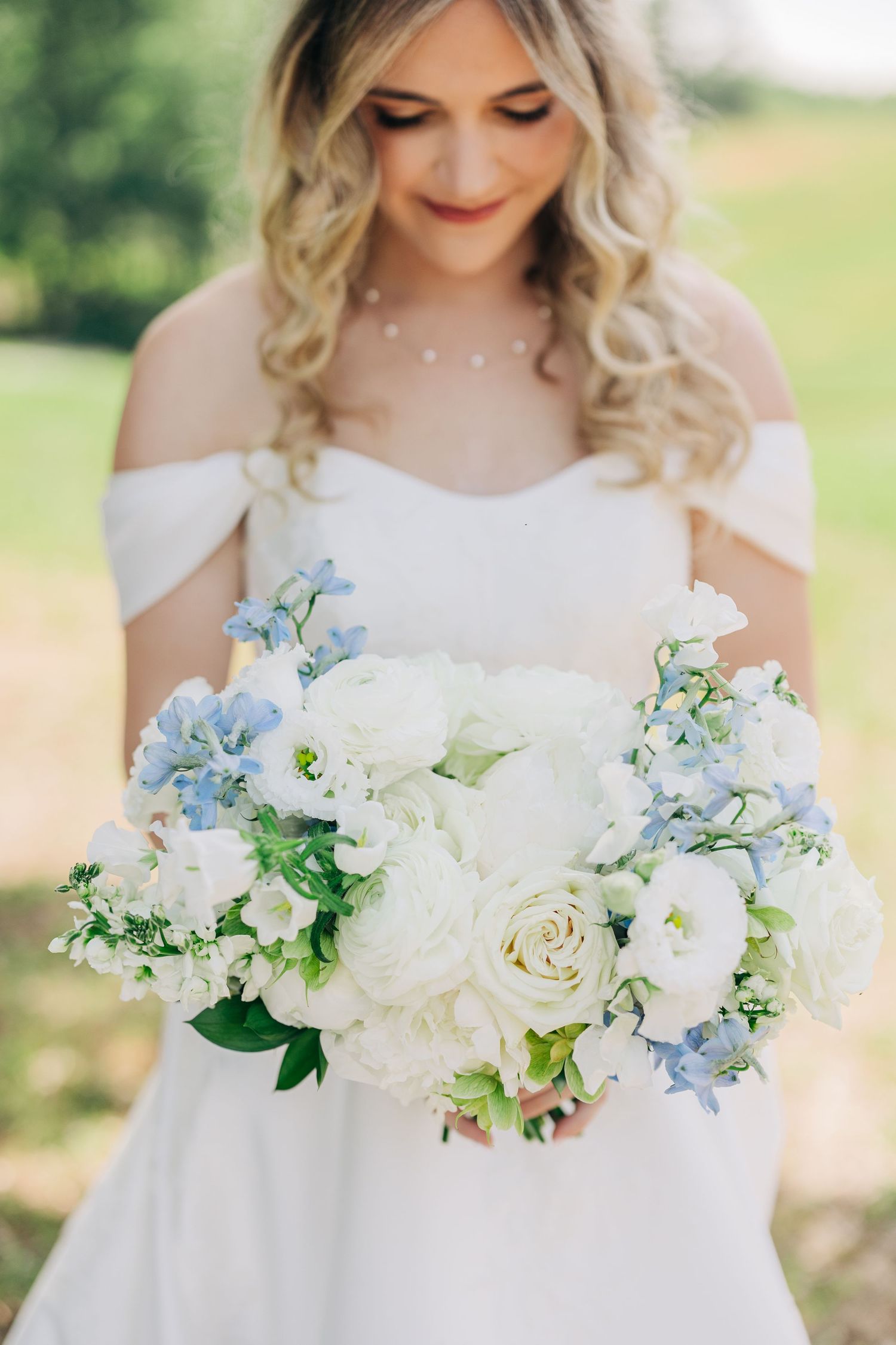 Bride in white off-shoulder gown holding large bouquet of white roses and soft blue flowers outdoors.