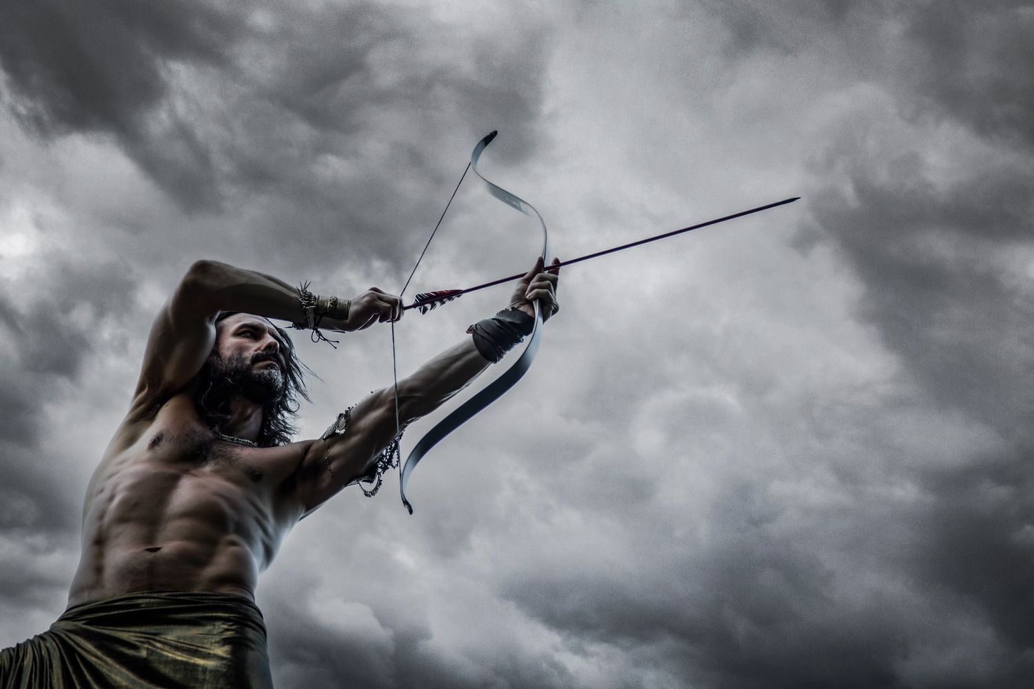 A muscular warrior with long hair draws a bow and arrow against a dramatic stormy sky.