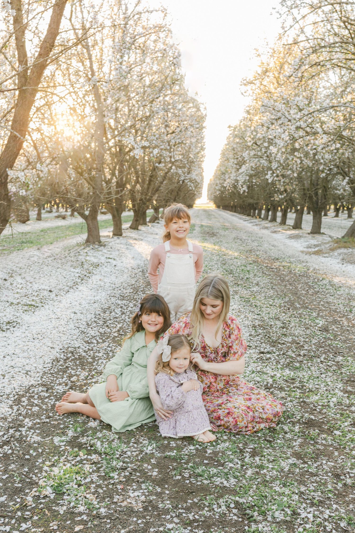 A mother and three daughters sit together among blooming trees with white petals on the ground in a serene orchard setting.