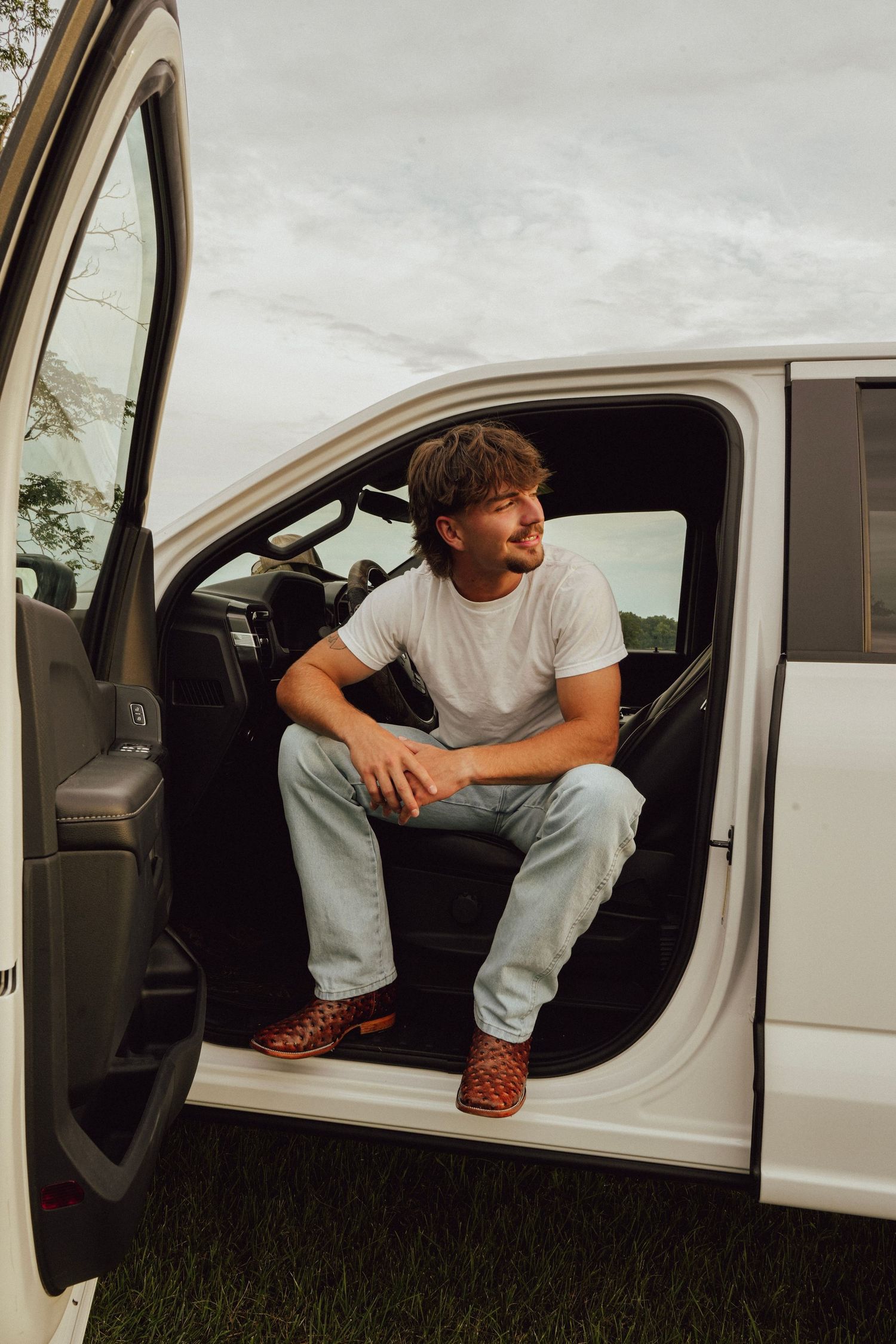 Young man in white t-shirt and jeans sitting in open door of white pickup truck, smiling outdoors.