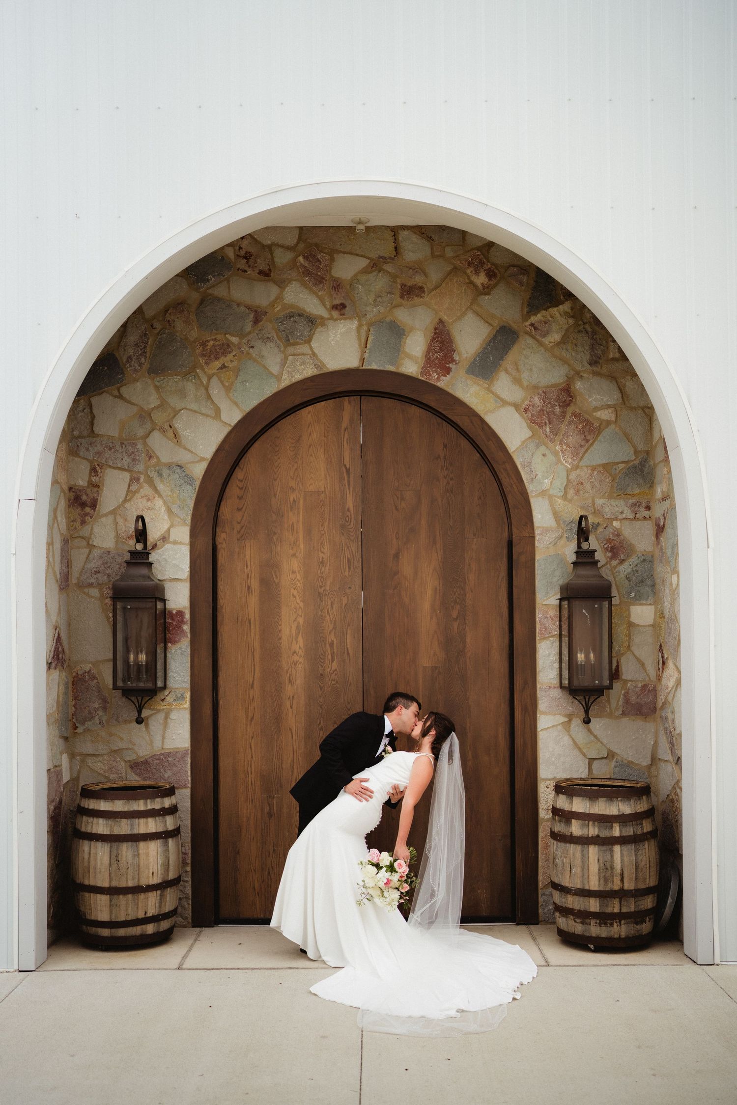 Bride and groom share a romantic dip kiss in front of a rustic arched wooden door with stone accents and wine barrels.