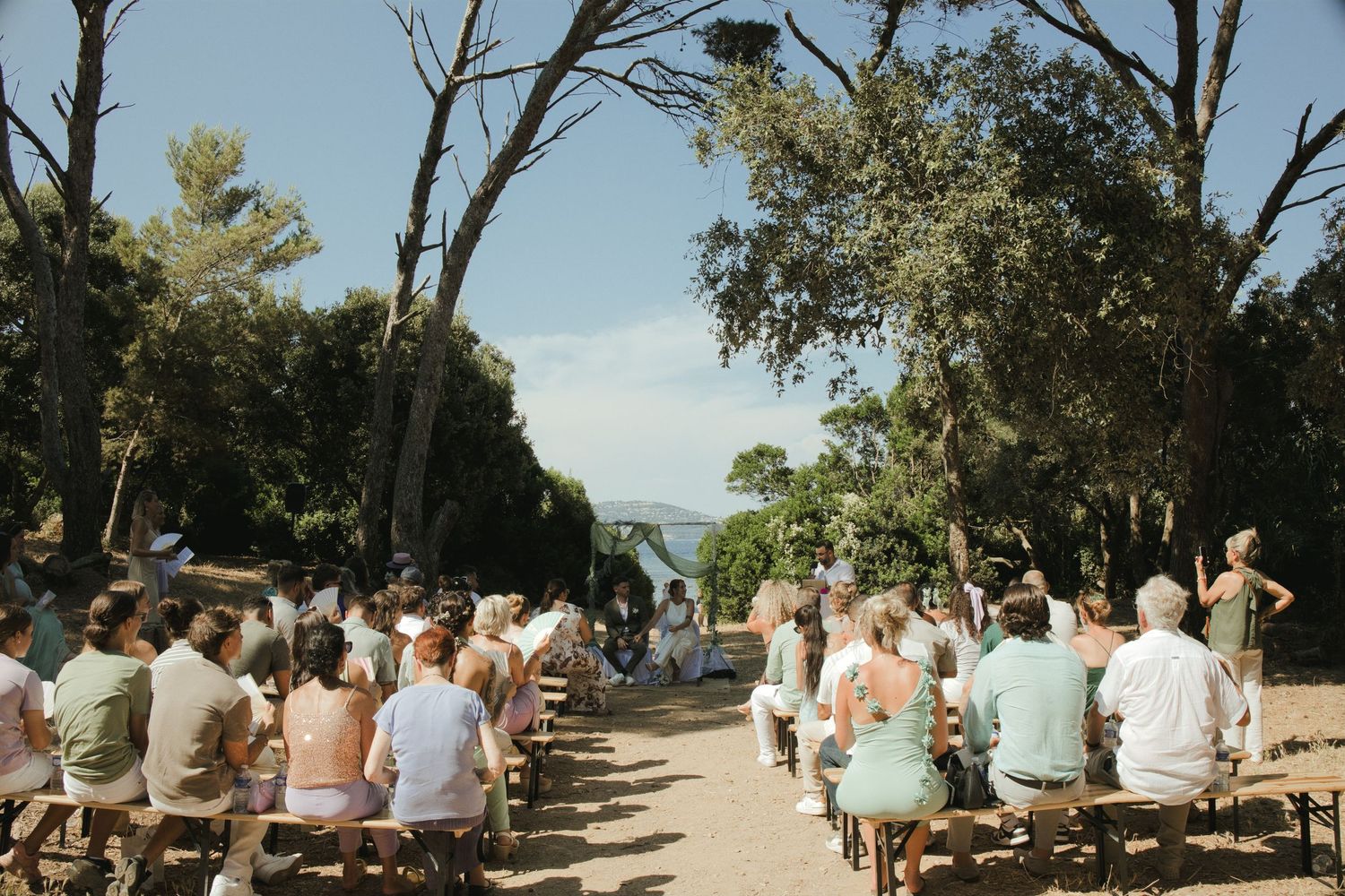 Vue d’ensemble d’une cérémonie de mariage sous les arbres, avec les invités de part et d’autre de l’allée.