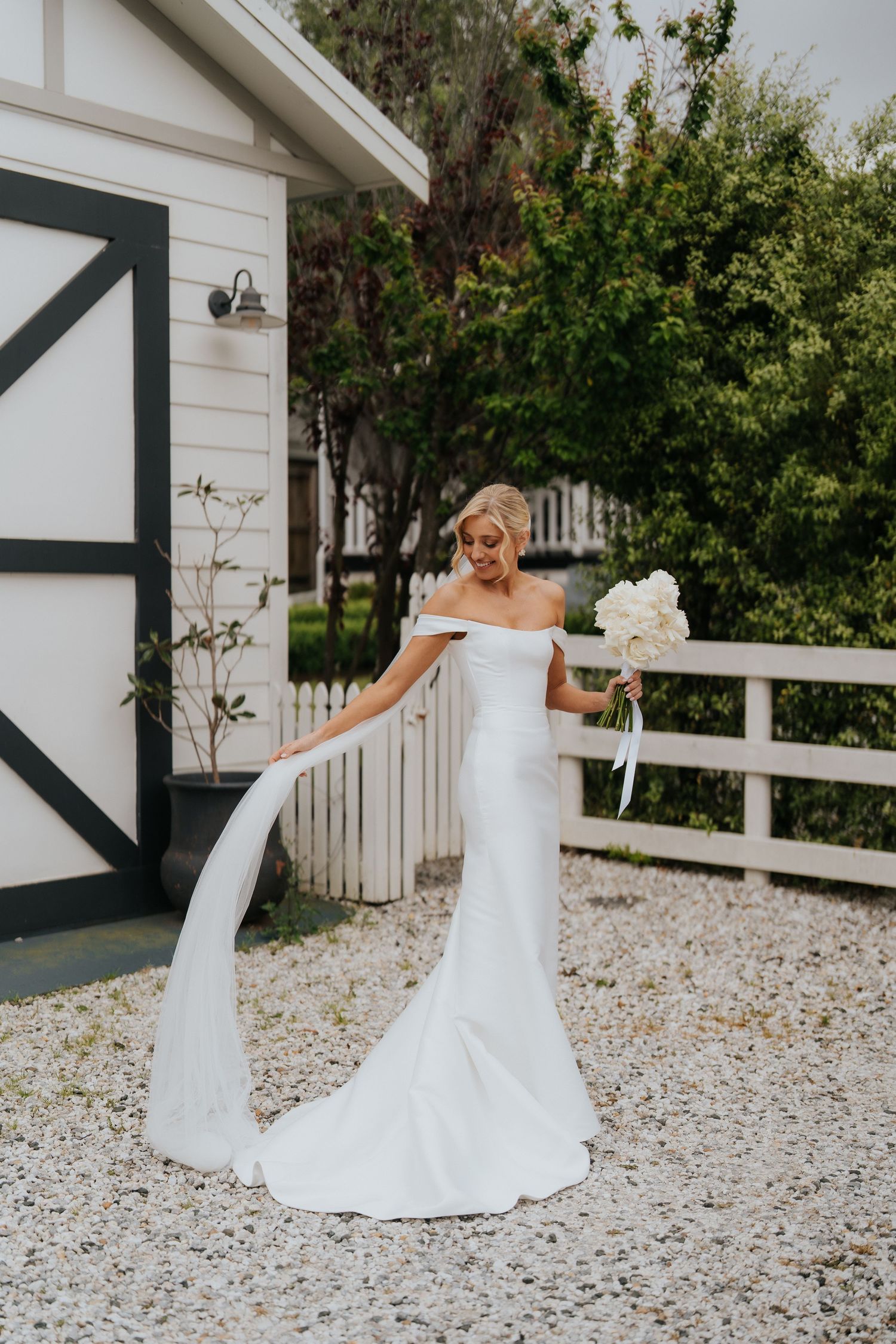 Bride in off-shoulder white mermaid gown holding bouquet of white flowers, standing on gravel path near white fence.