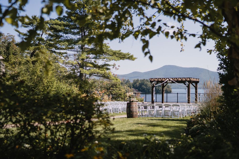 Outdoor wedding ceremony setup with white chairs and wooden pergola overlooking mountains and lake surrounded by lush greenery.