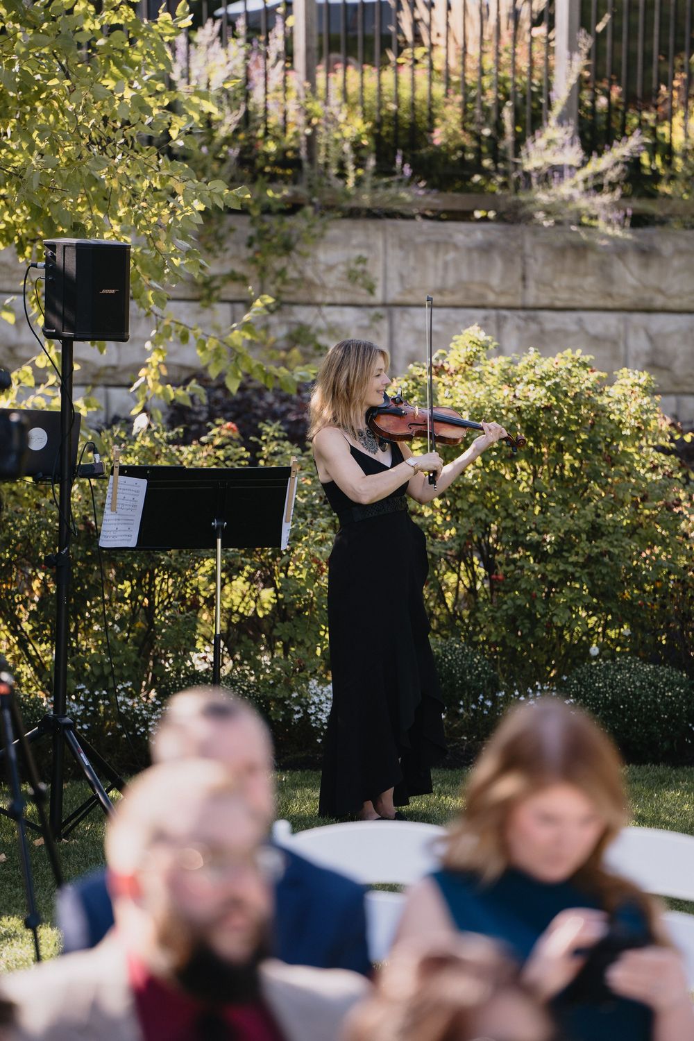 Woman in black dress playing violin outdoors at a garden event with audience seated in foreground.