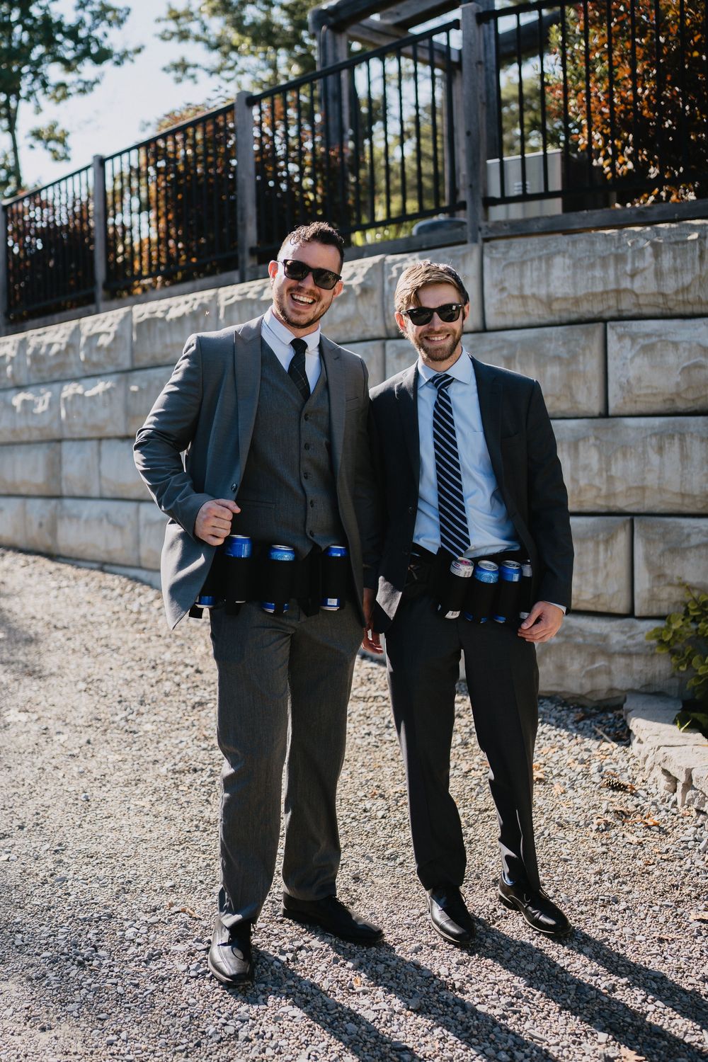 Two grooms in suits wearing beer can belts, smiling outdoors at a wedding celebration.