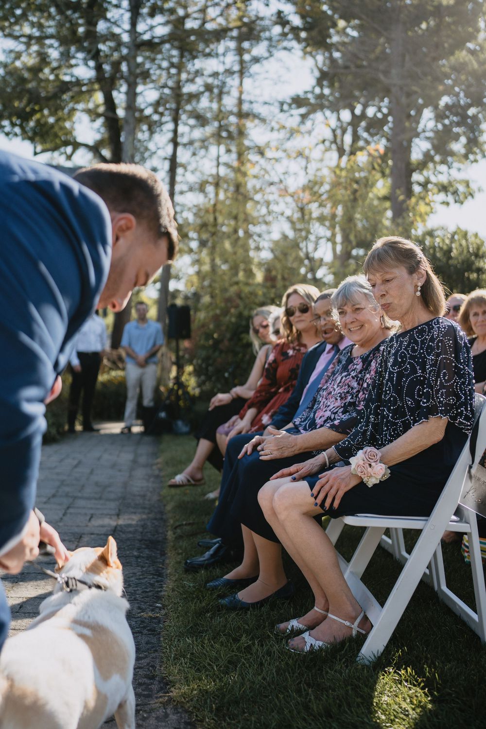 Guests smile at a dog during an outdoor wedding ceremony, seated on white chairs surrounded by lush greenery.
