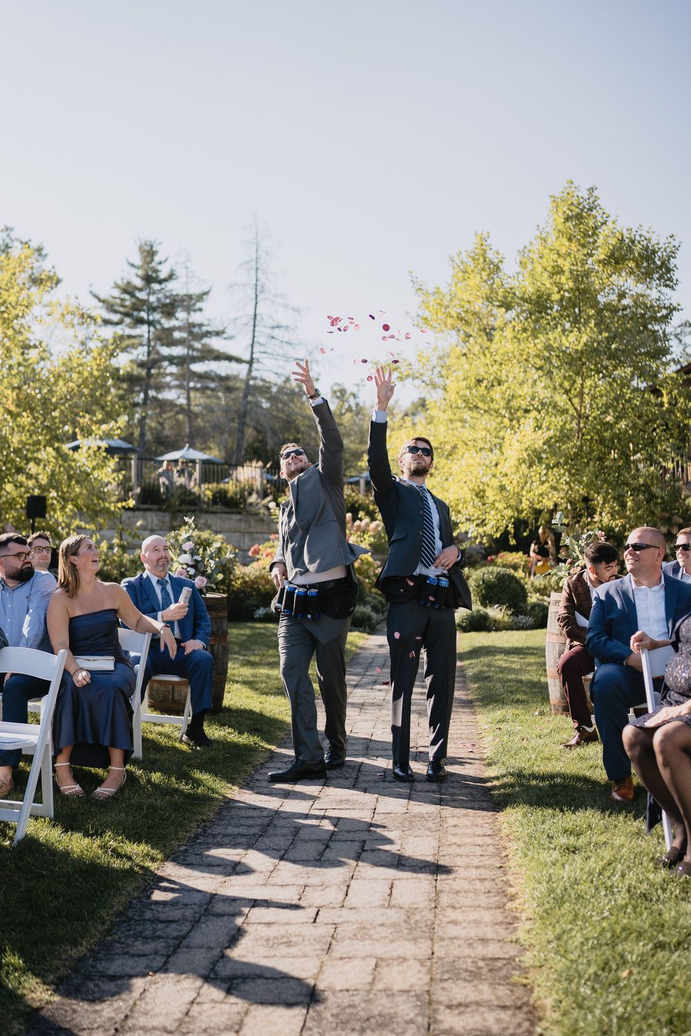 Two groomsmen toss rose petals in the aisle during an outdoor wedding ceremony with guests seated on white chairs.