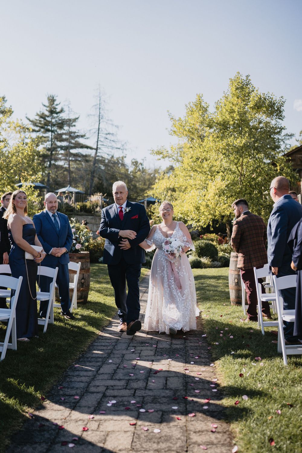 Bride walks down outdoor wedding aisle with escort, white chairs, rose petals, lush greenery, sunny day ceremony.
