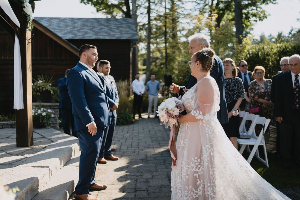 Bride in lace gown holding bouquet faces groom in navy suit during outdoor wedding ceremony on brick patio.
