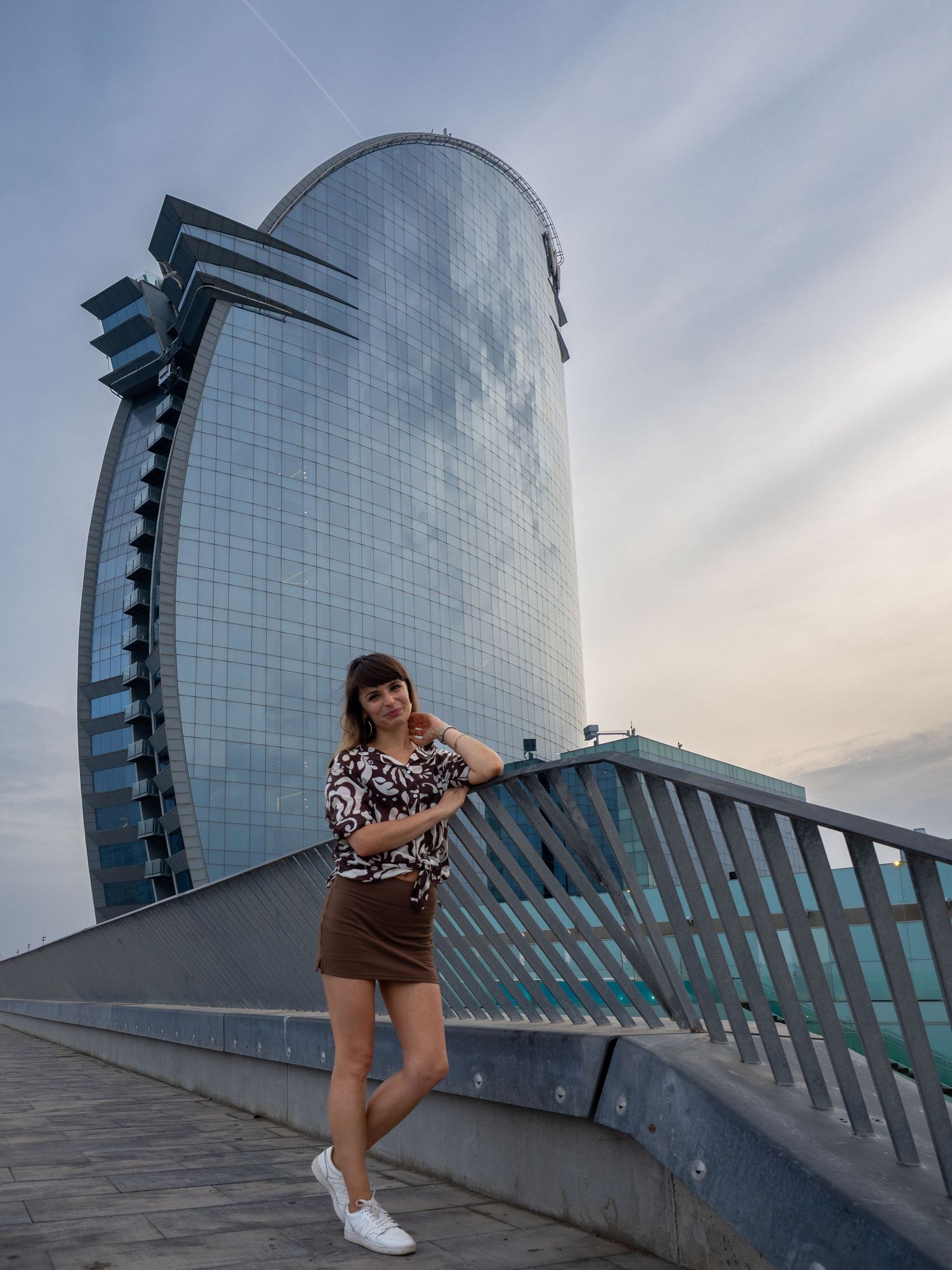 Woman posing in front of the W Barcelona hotel sail-shaped skyscraper on a cloudy day.
