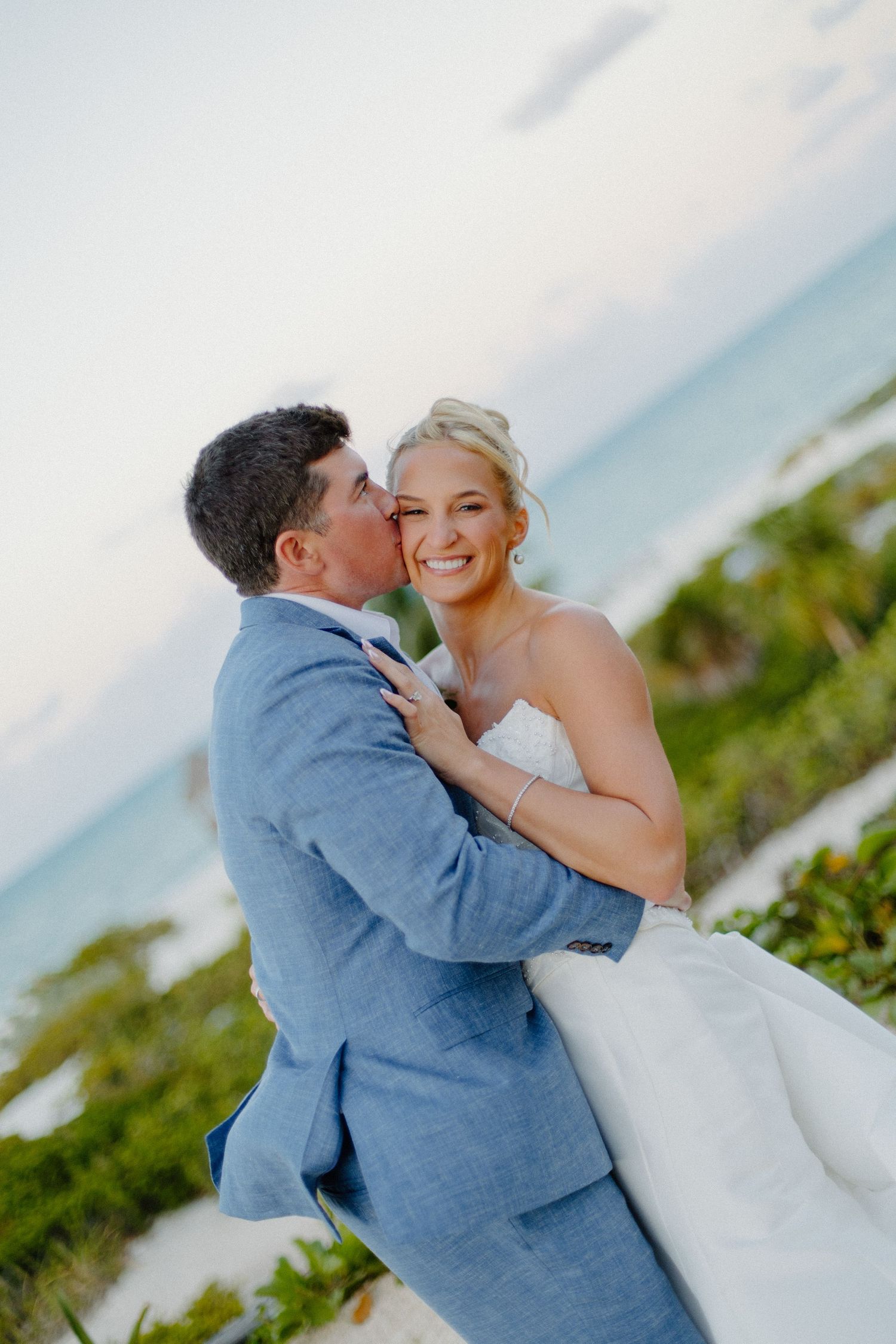 Groom in blue linen suit kisses smiling blonde bride on cheek with turquoise ocean and tropical foliage behind them.