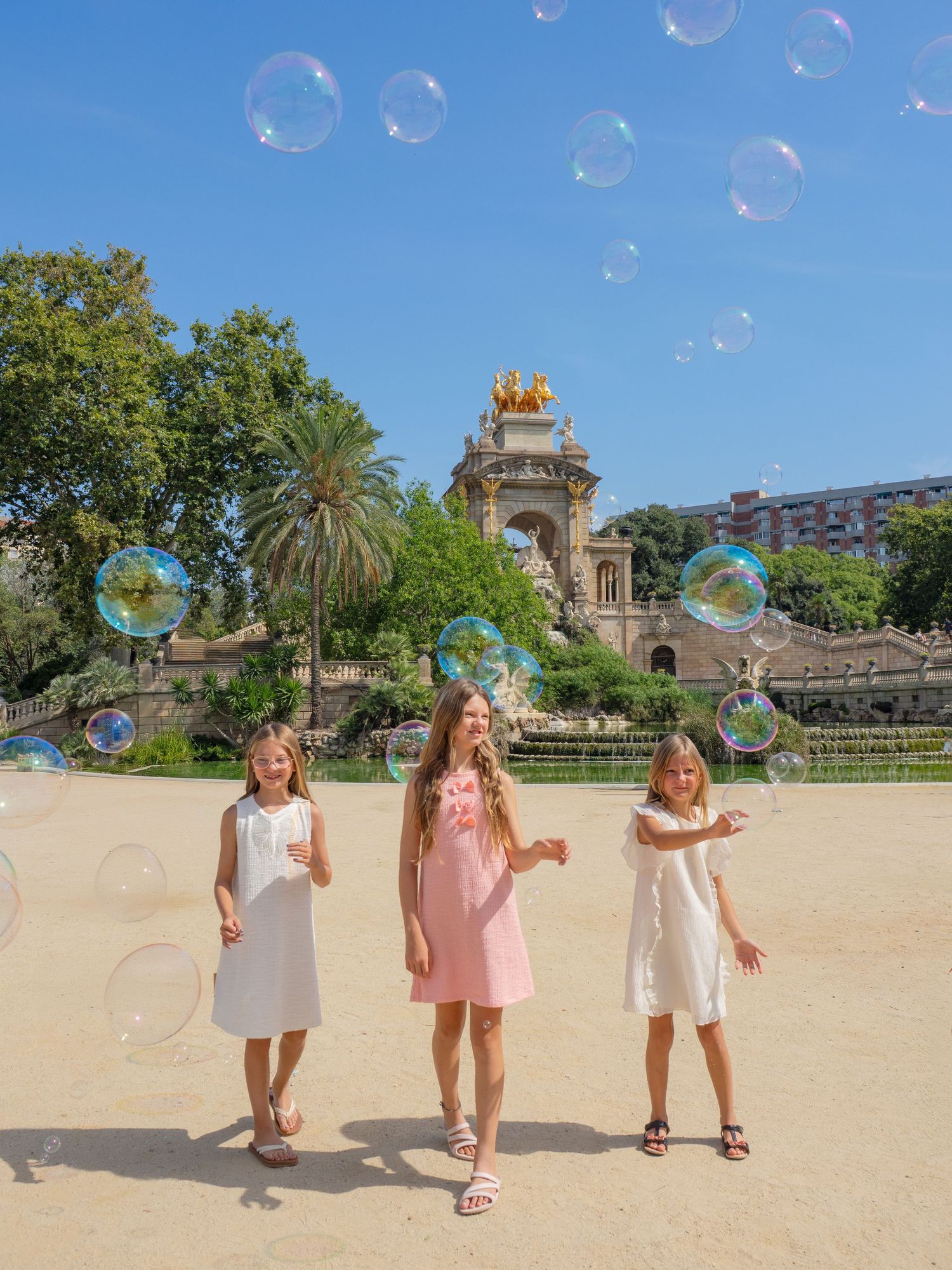 Three girls in dresses playing with bubbles in front of a golden fountain in Parc de la Ciutadella, Barcelona.