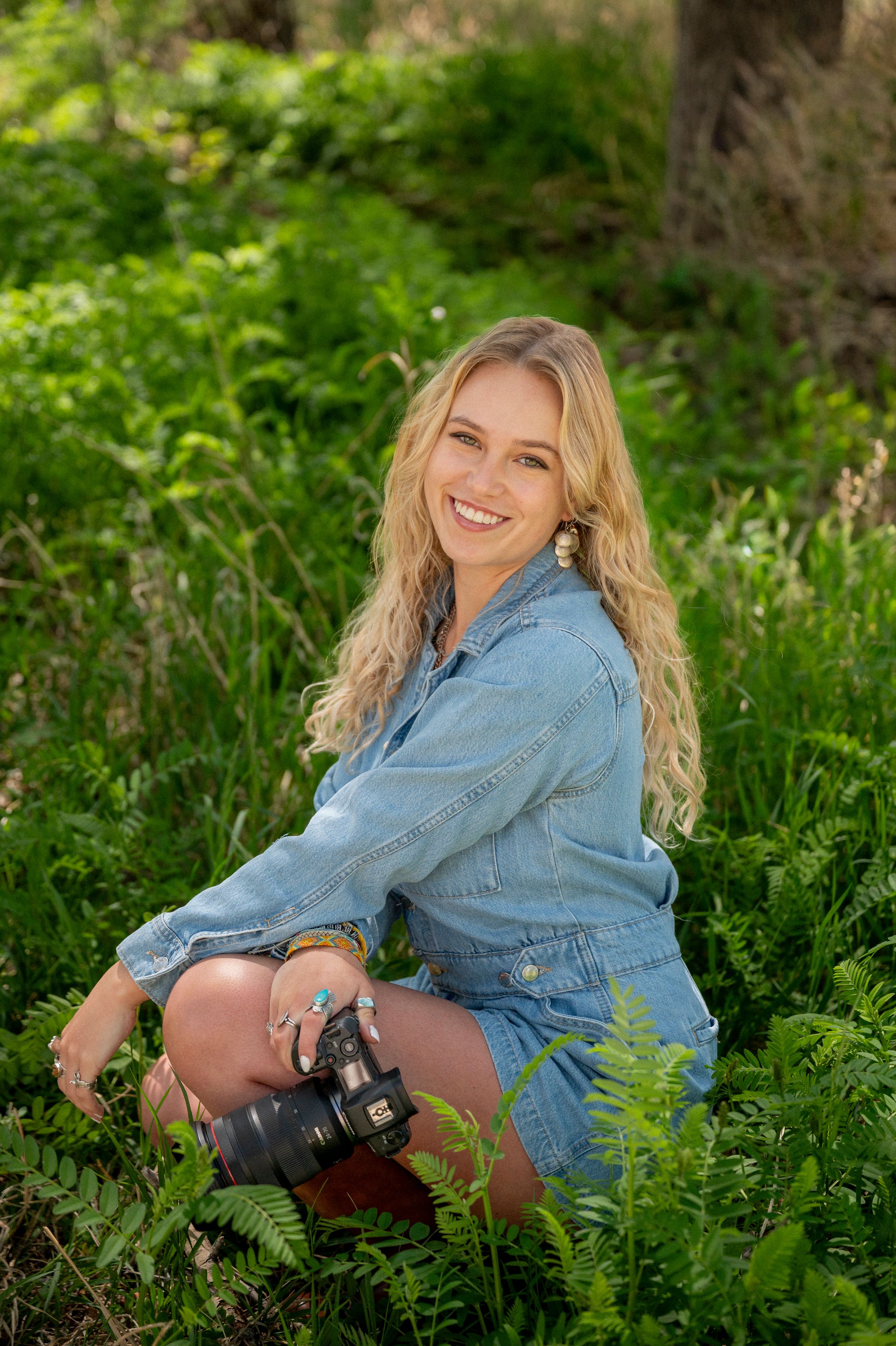 Smiling blonde woman in denim outfit sitting in lush green ferns holding a camera outdoors.