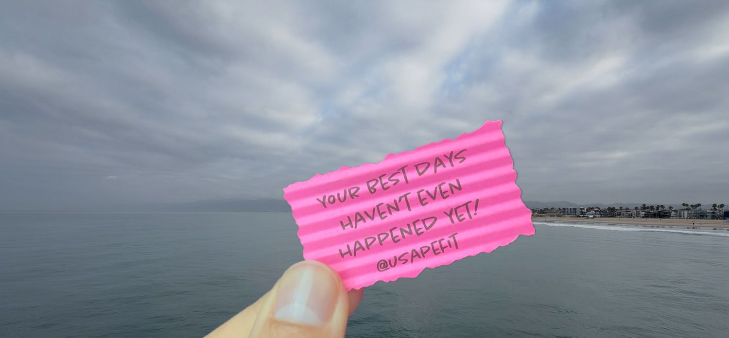 Hand holding a pink card with motivational quote against cloudy sky and ocean backdrop at the beach.