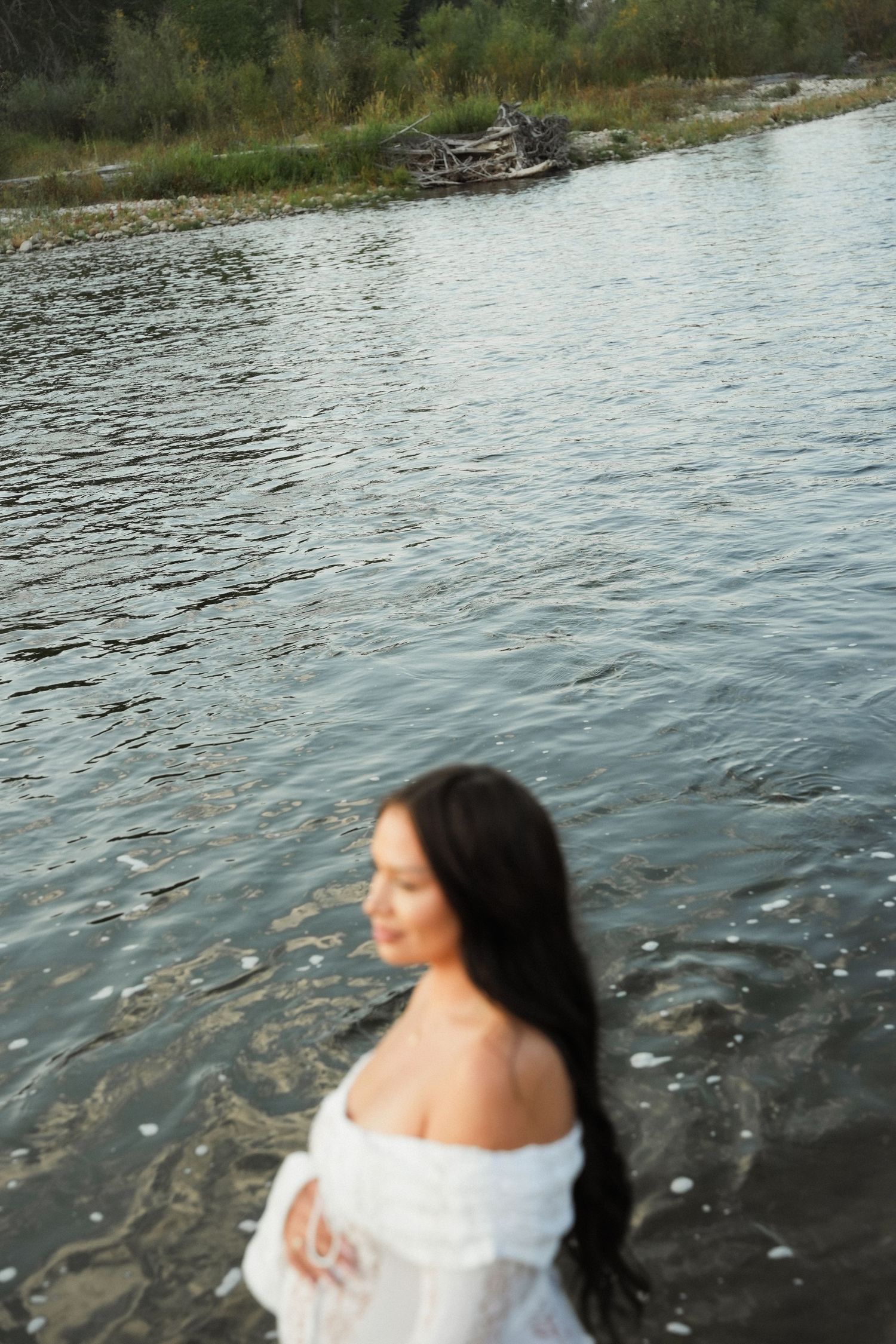 Woman in white off-shoulder dress standing in a calm river with rocky shores and driftwood in the background.