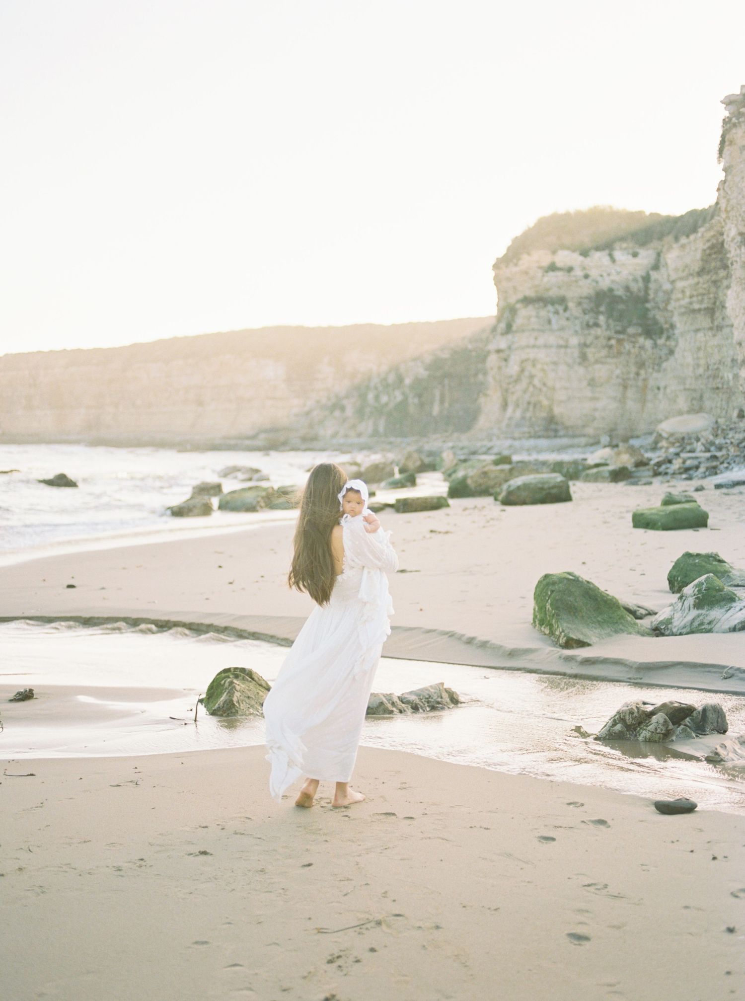 A mother in a white dress holds her newborn on a sunlit beach with rocky cliffs and golden light.