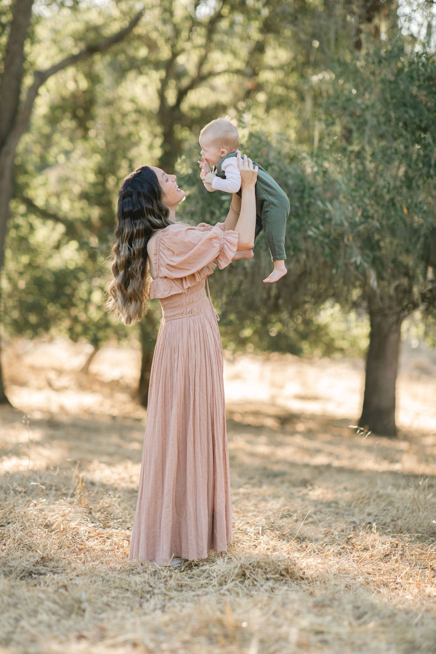 A family of four poses outdoors among trees, parents holding two young children, wearing neutral and earth-toned outfits.