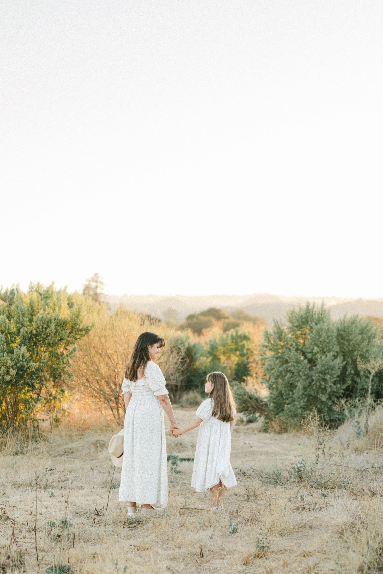 A mother and daughter in matching white dresses hold hands in a golden field at sunset.
