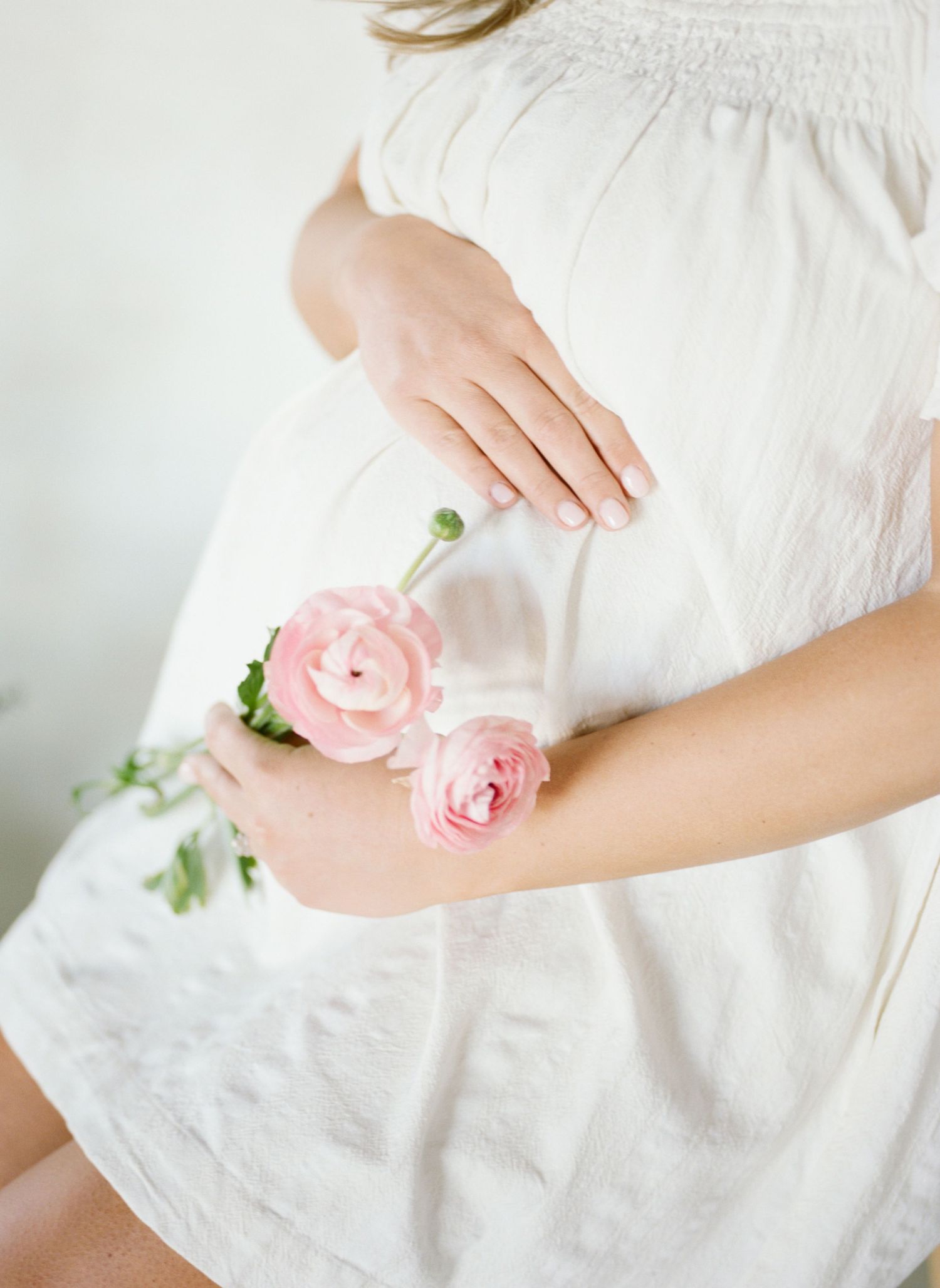 Pregnant woman in white dress holding pink ranunculus flowers against her baby bump in a soft, ethereal maternity photo.
