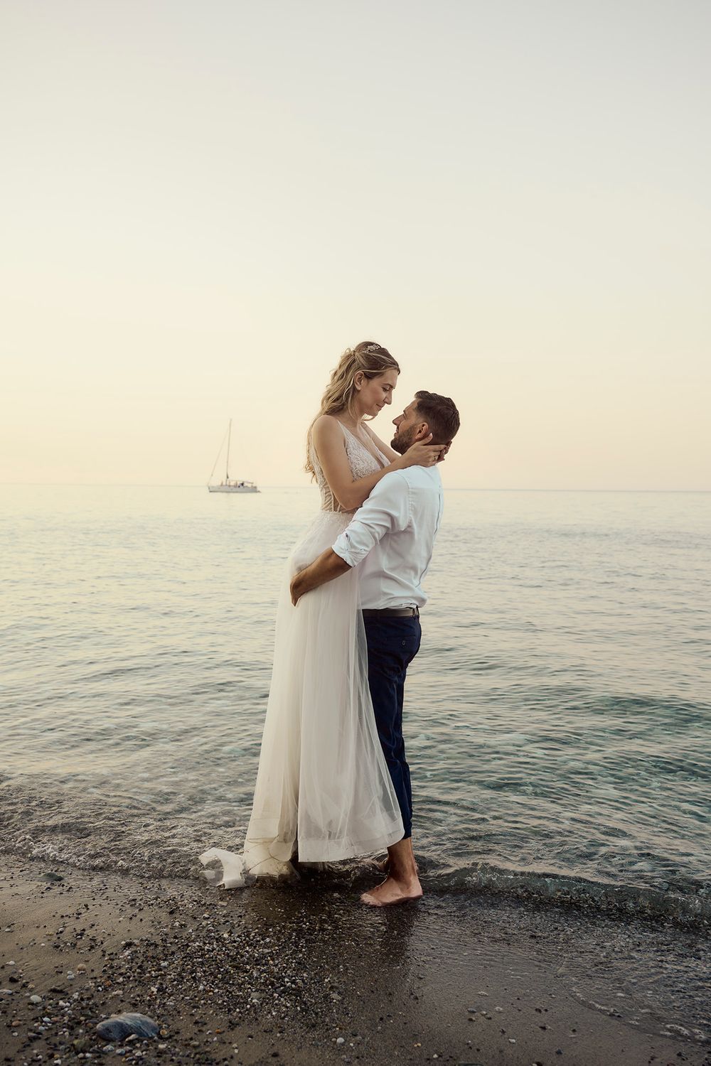 Romantic couple in white embracing on a sandy beach with a sailboat at sunset.