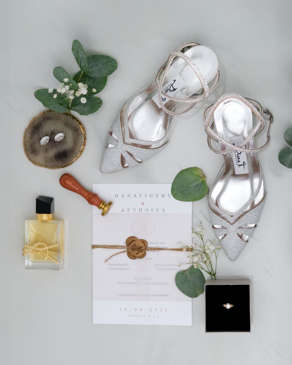 Flat lay of wedding bridal details including silver shoes, perfume, invitation, and rings.