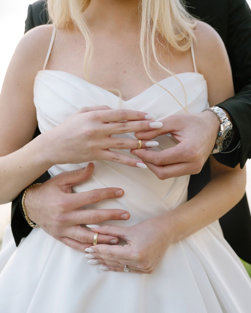 Close-up of groom embracing bride from behind, showcasing gold wedding rings.