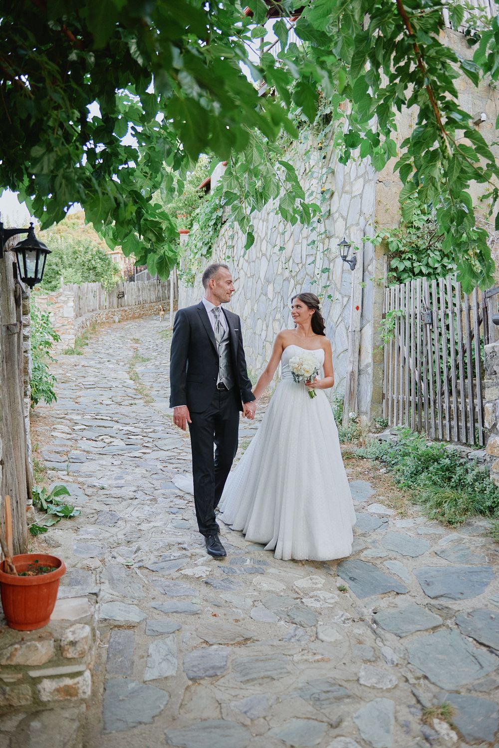 Bride and groom walking hand-in-hand down a traditional stone village path in Greece.
