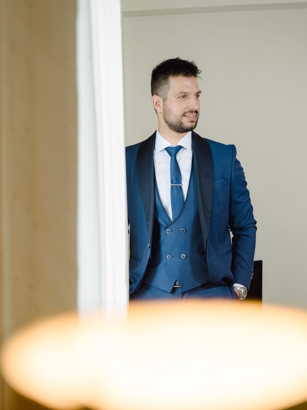 Groom in a blue three-piece suit looking out a window during wedding prep.